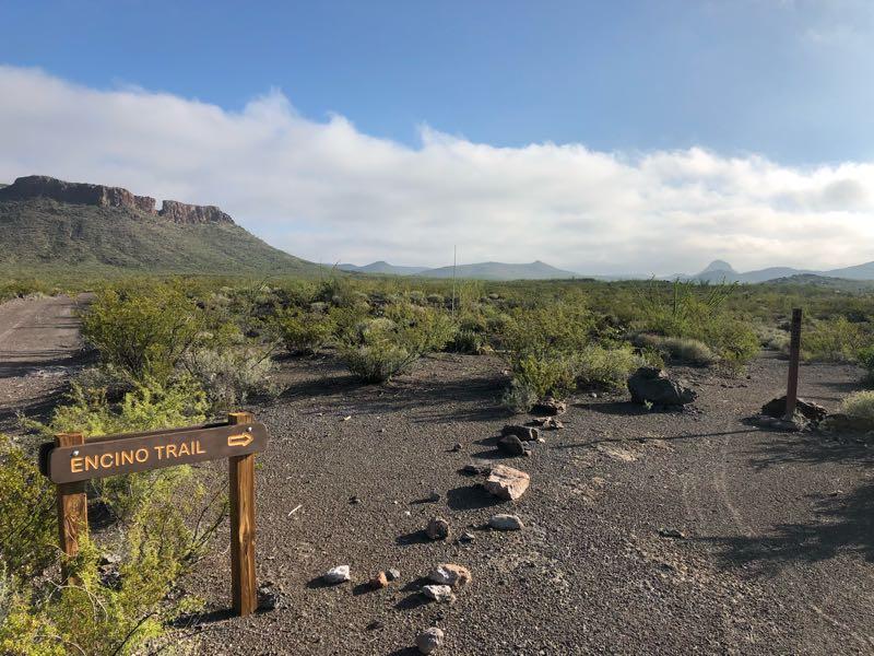 A dirt trail sign labeled "Encino Trail" points to the right, surrounded by a rocky landscape with sparse vegetation. In the background, mountains rise under a partly cloudy sky. Big Bend State Park mountain bike trail.
