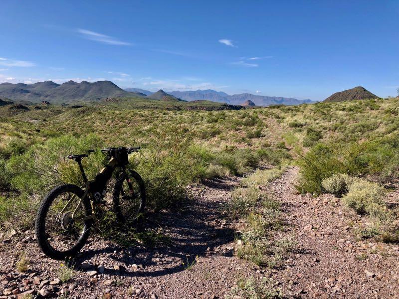 A mountain bike leaning against a bush on a rocky trail, surrounded by sparse vegetation and rolling hills under a clear blue sky. In the background, mountains stretch across the horizon. Big Bend State Park mountain bike trail.