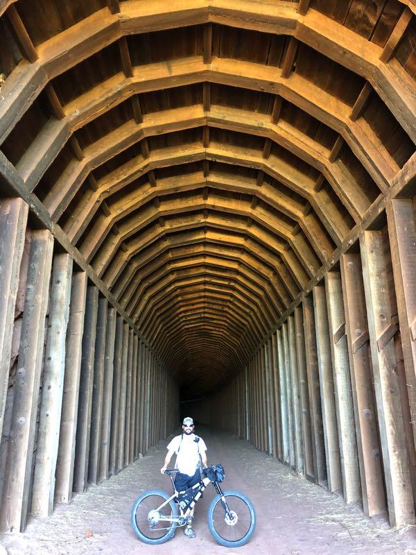 A person stands next to a mountain bike inside a large wooden structure with a high arched roof. The interior features a series of wooden beams and vertical supports, creating a tunnel-like effect leading into darkness. Sunlight illuminates the entrance, highlighting the texture of the wood and the sandy floor. Caprock Canyons Trailway mountain bike trail.