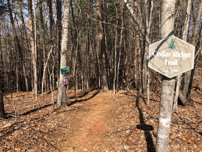 A dirt trail leads into a wooded area, flanked by tall trees with bare branches and scattered fallen leaves on the ground. Two signs are visible: one on the left providing information about the trail, and a hexagonal sign on the right that reads "Cedar Ridge Trail, Est. 1999." The scene captures a serene and inviting atmosphere typical of a nature trail. Cedar Ridge Chatmoss mountain bike trail.