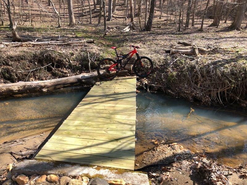 A mountain bike rests on a wooden bridge that crosses a shallow creek in a wooded area. The ground is covered with fallen leaves, and trees can be seen in the background, indicating a natural, outdoor setting. Cedar Ridge Chatmoss mountain bike trail.