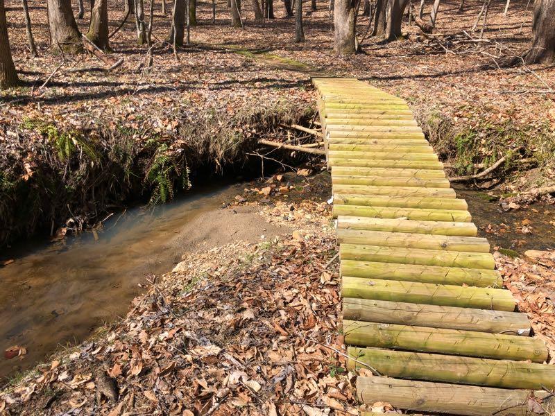 A wooden footbridge crossing a narrow stream in a wooded area, surrounded by fallen leaves and trees. The bridge is made of logs laid side by side, providing a natural pathway over the water. Cedar Ridge Chatmoss mountain bike trail.
