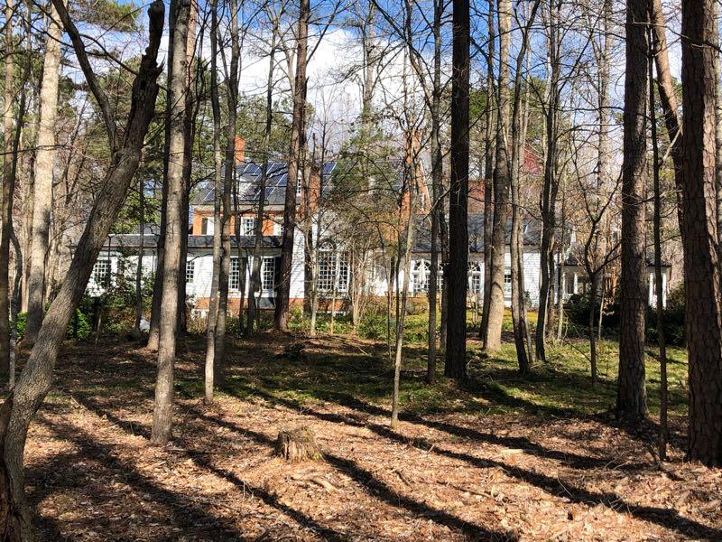 A large, two-story house partially obscured by trees, featuring a combination of brick and white siding. Solar panels are visible on the roof. The surrounding area is filled with tall trees and a carpet of fallen leaves, creating a serene, natural setting. Sunlight filters through the branches, illuminating the landscape. Cedar Ridge Chatmoss mountain bike trail.