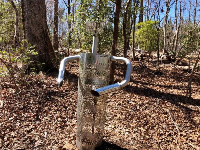 A metallic sculpture resembling a humanoid figure stands in a wooded area, surrounded by fallen leaves and trees. The sculpture features a perforated cylindrical body, two arms made from shiny pipes, and an abstract head. Sunlight filters through the trees, casting dappled light on the scene. Governor's Creek mountain bike trail.