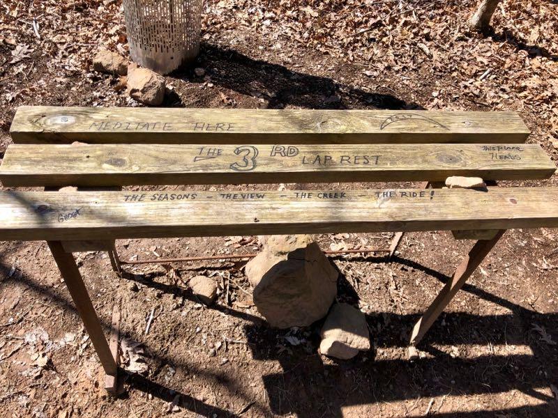 A wooden bench in a natural outdoor setting, featuring engraved messages that include "Meditate Here," "The 3rd Lap Rest," and "The Seasons, The View, The Creek, The Ride!" The bench is supported by wooden legs and surrounded by fallen leaves and rocks. Governor's Creek mountain bike trail.