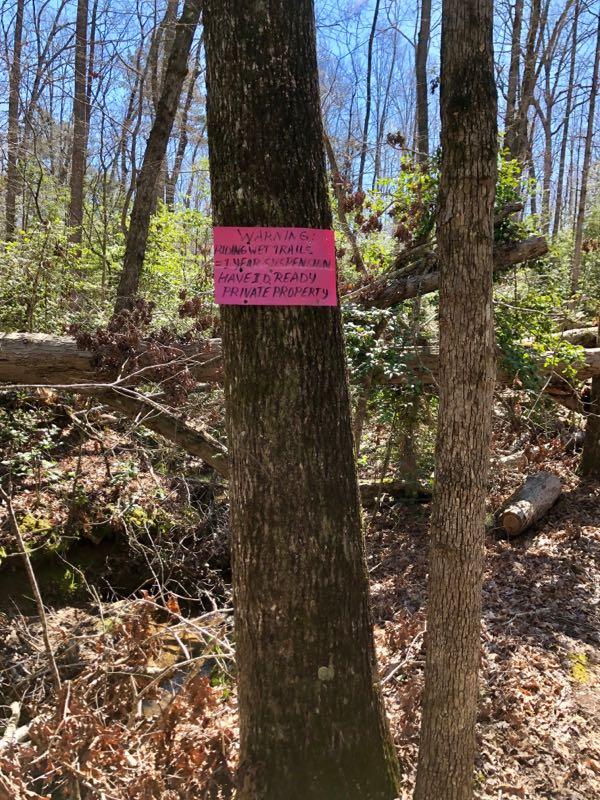 A bright pink warning sign attached to a tree in a wooded area, stating "Warning: Swimming Trail - Private Property." Surrounding the tree are other trees and some fallen branches, indicating a natural outdoor setting. Governor's Creek mountain bike trail.