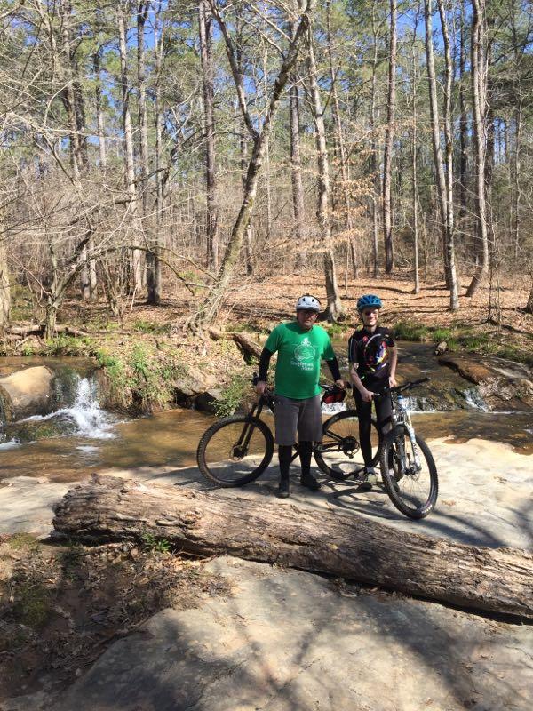 Two mountain bikers stand next to their bicycles on a rocky area near a creek, surrounded by a wooded landscape. The scene is illuminated by sunlight filtering through the trees. One rider wears a green shirt and helmet, while the other is dressed in a colorful cycling outfit and a blue helmet. A small waterfall can be seen in the background. Hard Labor Creek State Park mountain bike trail.