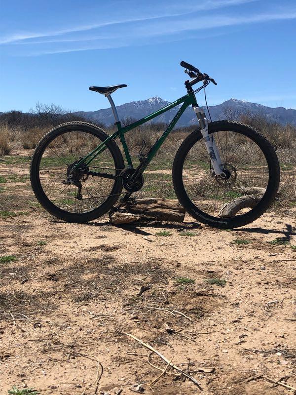 A green mountain bike is parked on sandy ground with a mountainous landscape in the background. The sky is clear with a few wispy clouds, and the scene is surrounded by sparse vegetation. San Pedro Riparian National Conservation Area mountain bike trail.