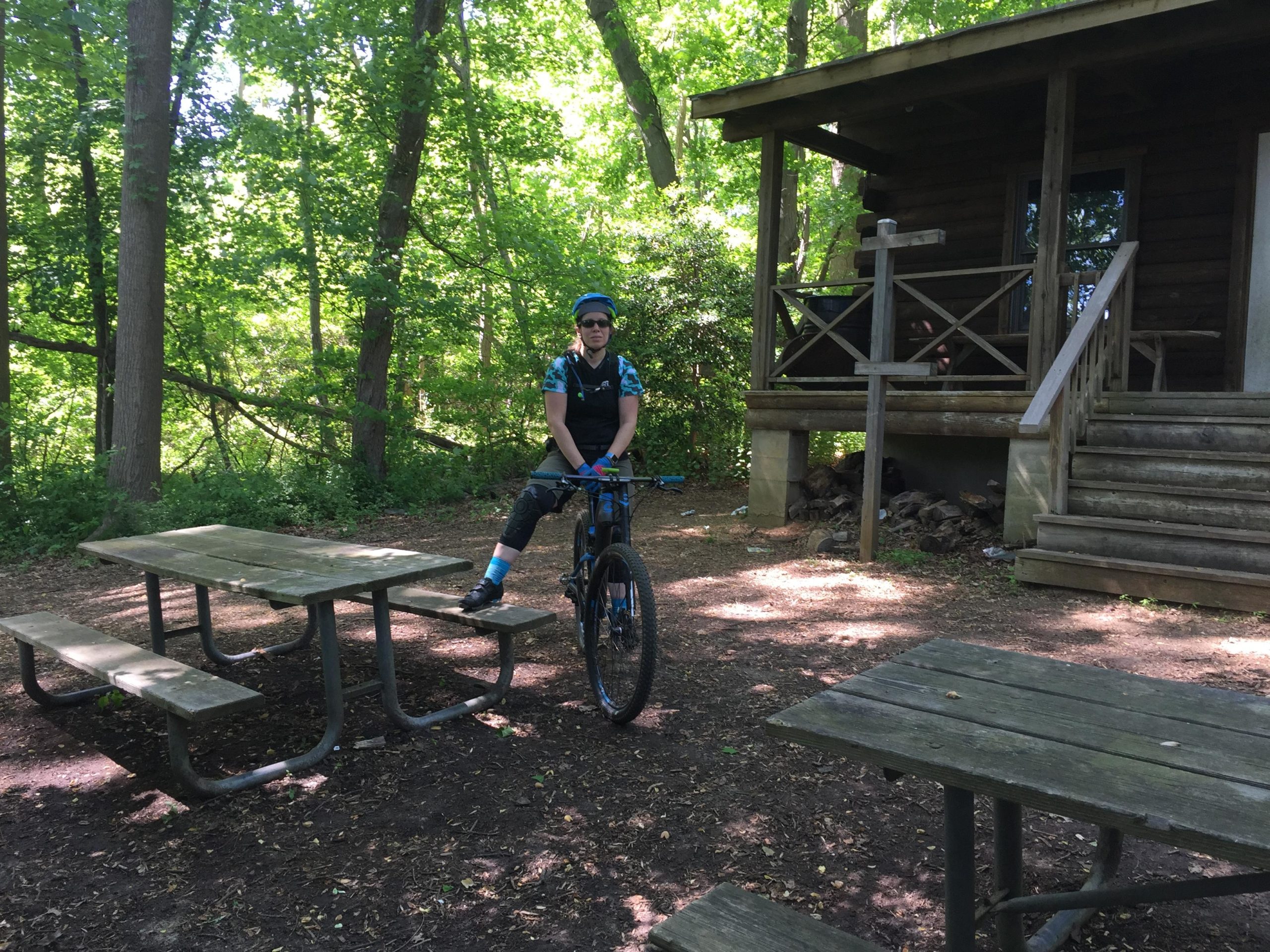 A person sitting on a mountain bike next to a wooden picnic table in a wooded area, with a cabin partially visible in the background. The scene is set in a green, natural environment with sunlight filtering through the trees. Hartshorne Woods Park mountain bike trail.