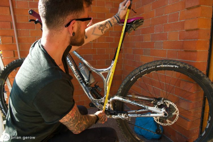 A person measuring the frame of a mountain bike with a tape measure, positioned near a brick wall. The individual has visible tattoos and is wearing sunglasses, while a backpack is visible in the background.