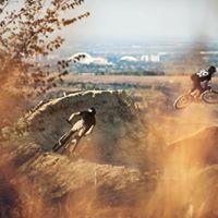 Two mountain bikers are riding on a dirt trail, one navigating a curved section while the other is slightly elevated, surrounded by tall grass and a distant landscape. Bukovac Bike Park mountain bike trail.