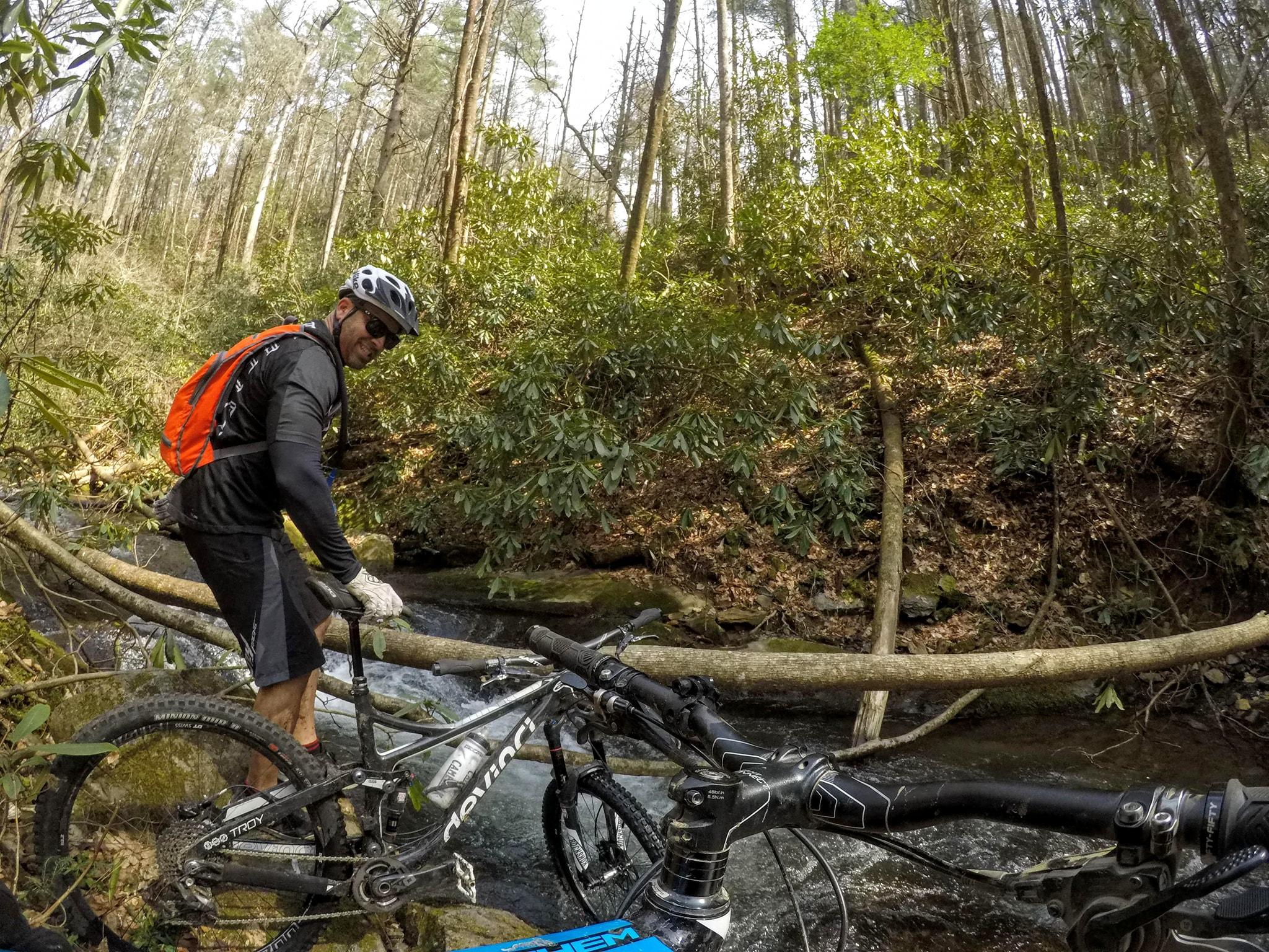 A mountain biker navigating a stream crossing in a forested area, wearing a helmet and an orange backpack. The bike is partially submerged in water as the rider balances on a log over the stream. Lush greenery surrounds the scene, with tall trees in the background and sunlight filtering through the leaves. Pinhoti Trail: Mountaintown Creek Segment mountain bike trail.