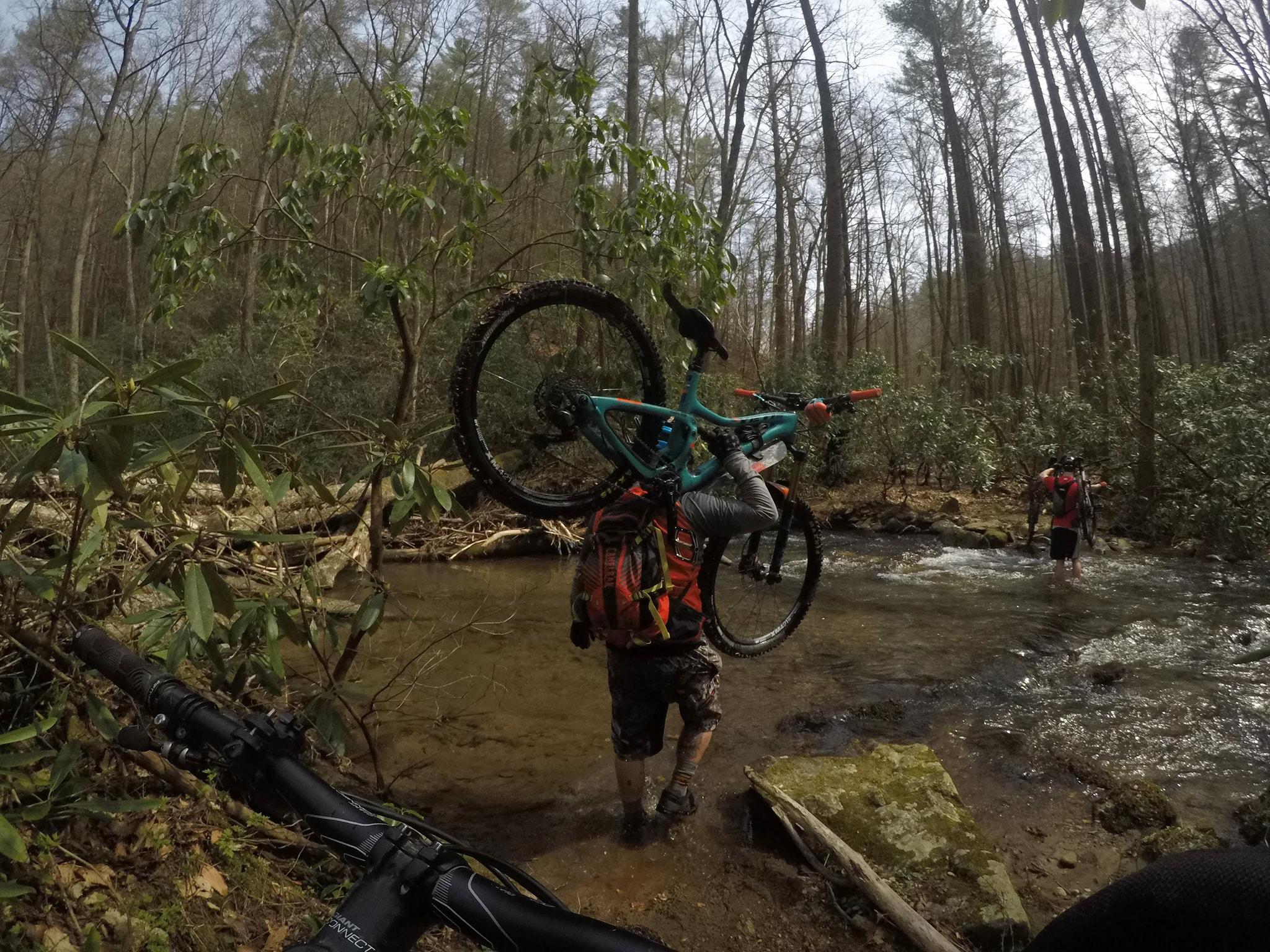 Two mountain bikers navigating a shallow stream in a forested area. One biker is holding their bike above their head while crossing the water, and the other is walking through the stream, carrying their bike. Lush greenery and tall trees surround the scene, indicating a remote outdoor setting. Pinhoti Trail: Mountaintown Creek Segment mountain bike trail.