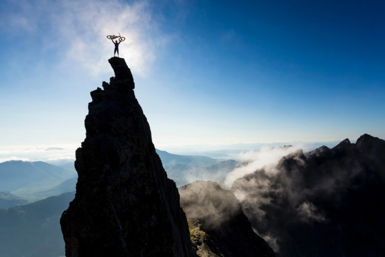 A silhouette of a person standing atop a tall rock pinnacle, raising a bicycle above their head, against a clear blue sky. Below, a vast landscape of mountains and valleys is shrouded in mist, creating a dramatic backdrop that captures the sense of achievement and adventure.