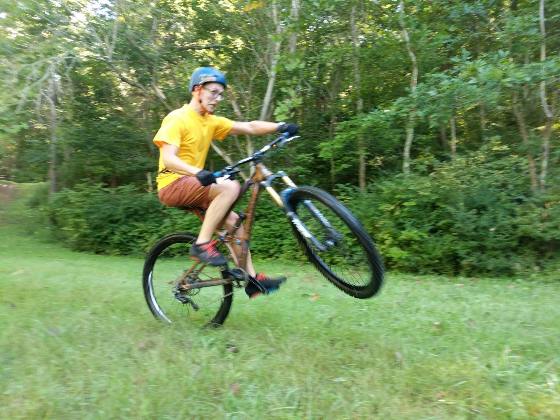Transition Covert: A person riding a mountain bike on a grassy trail, performing a wheelie. The cyclist is wearing a yellow shirt, brown shorts, and a blue helmet, surrounded by trees in a natural setting.