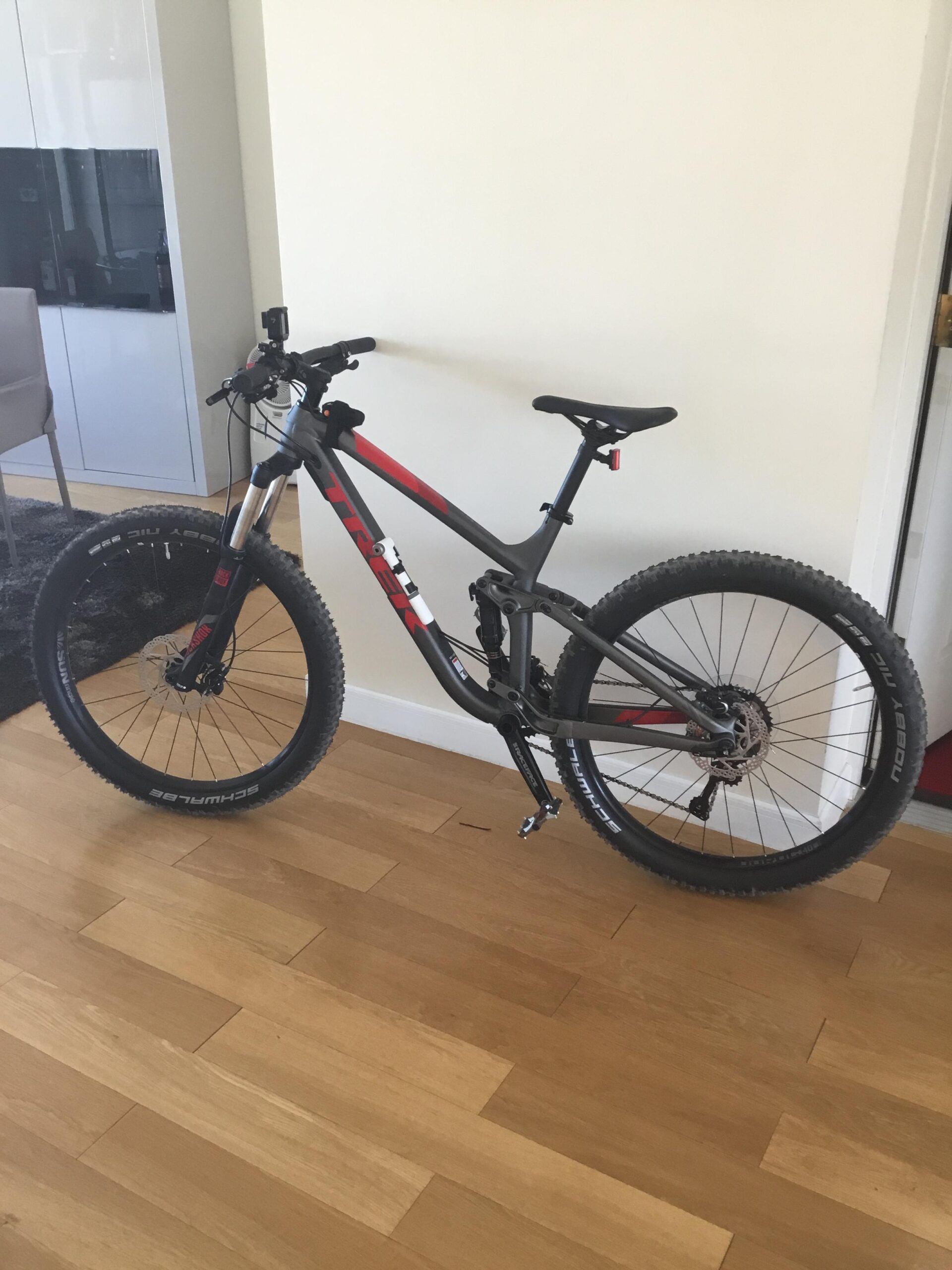 Trek Fuel EX 5: A mountain bike with a gray and red frame is leaning against a wall in a room with wooden flooring. The bike features large tires and a visible gear mechanism. In the background, there is a modern chair and furniture that suggest a contemporary interior design.
