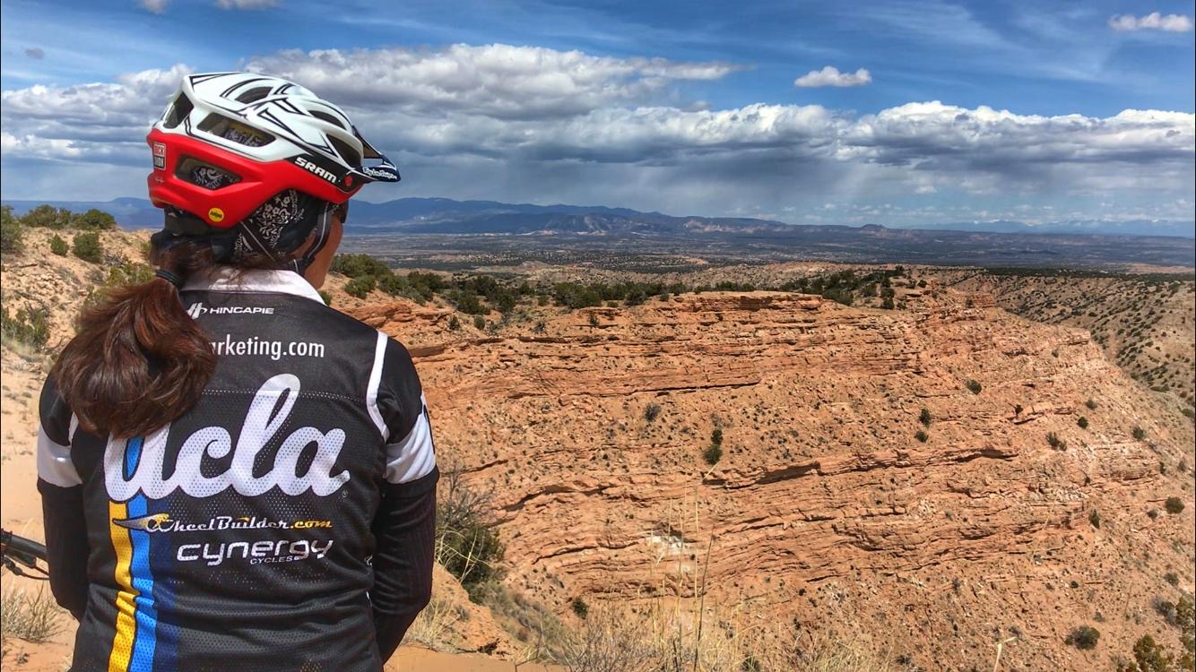 A person wearing a cycling jersey with "UCLA" printed on the back stands on a rocky hillside, looking out over a vast landscape of red rock formations and distant mountains under a partly cloudy sky. Mariposa Fat Bike Trails mountain bike trail.