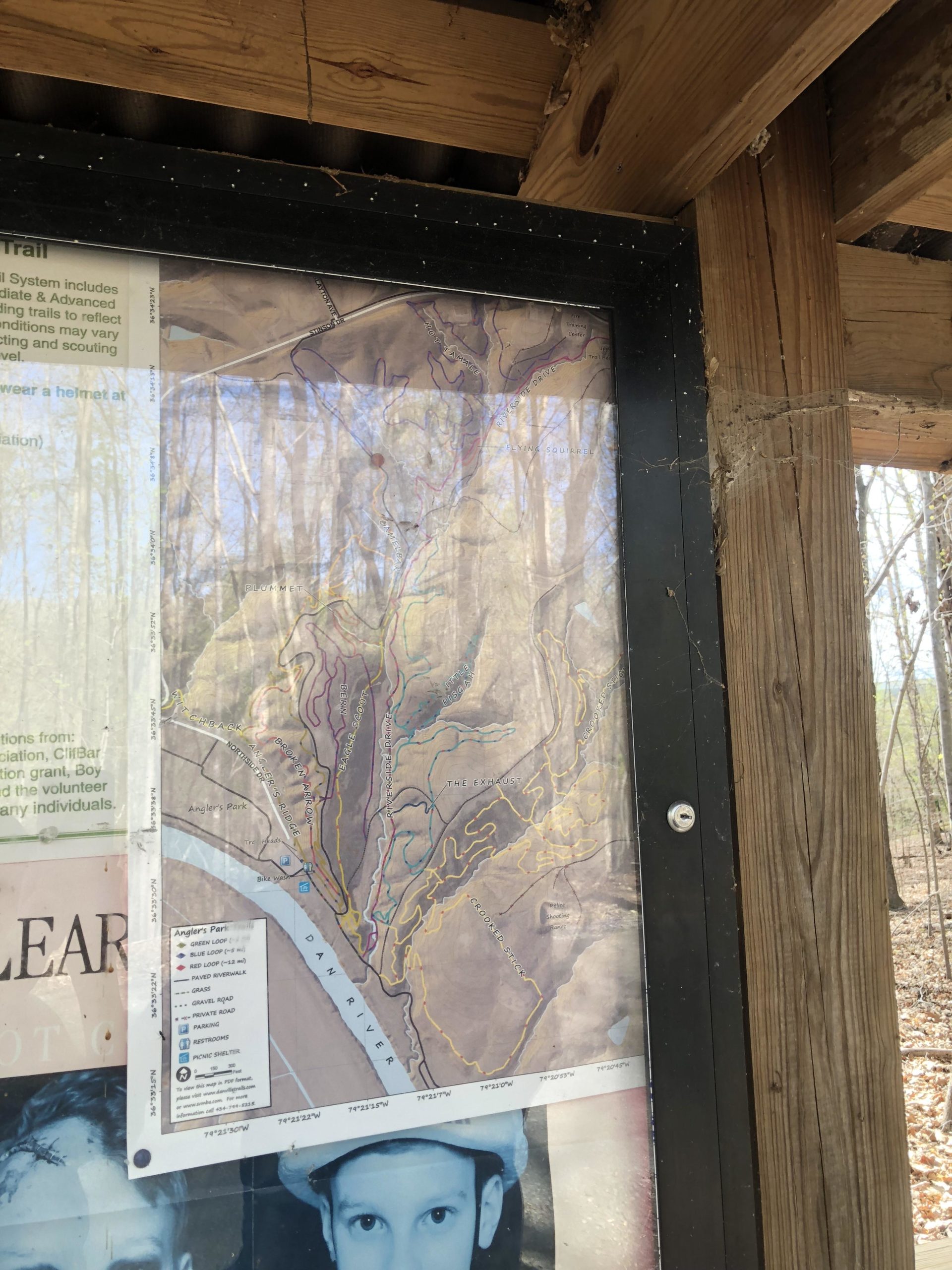 Map of a trail system displaying various routes and features along the Dan River, located on a wooden signpost in a wooded area. The map includes colored lines indicating different trails and nearby landmarks, alongside informational text about trail conditions and safety. Angler's Ridge mountain bike trail.
