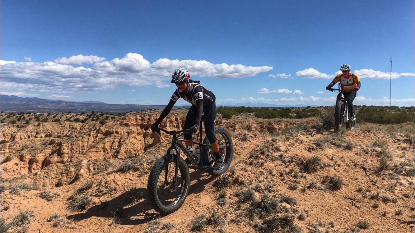 Two mountain bikers navigate a rugged, sandy trail with rocky terrain and low vegetation. The landscape features canyons and distant hills under a blue sky with scattered clouds. The cyclist in the foreground is focused on riding their bike, while the second cyclist trails behind. Mariposa Fat Bike Trails mountain bike trail.