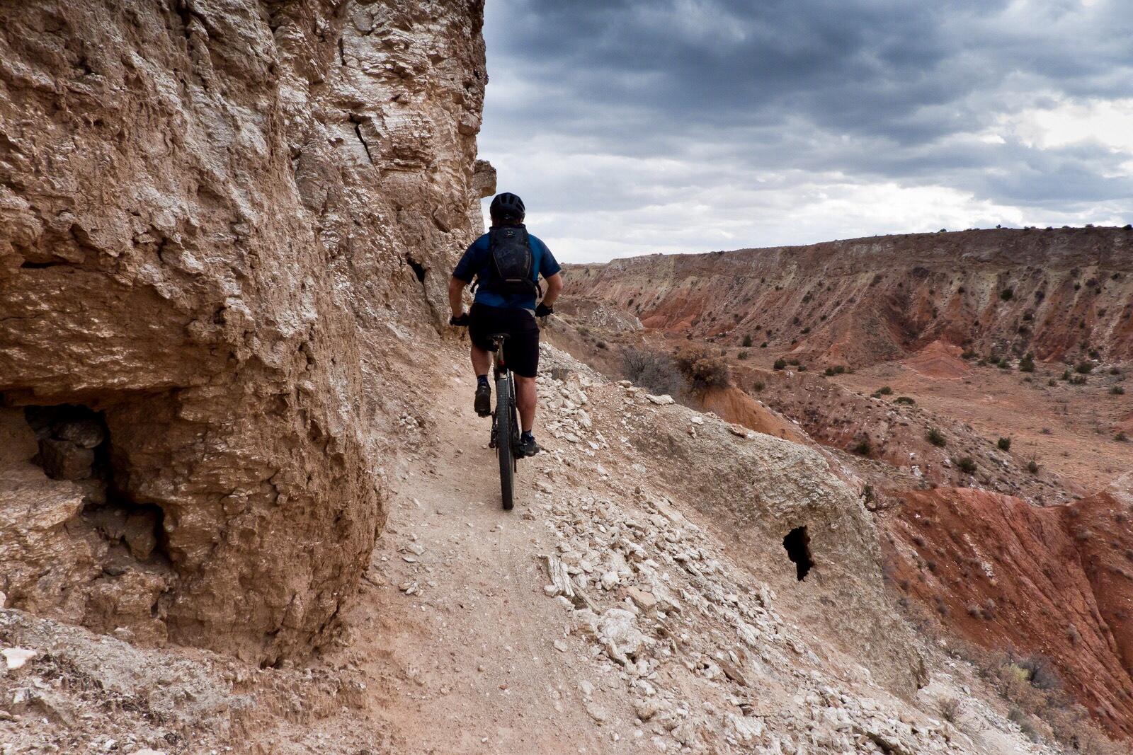 A mountain biker navigates a narrow dirt path along a rocky cliff, with a dramatic landscape of reddish-brown hills and a cloudy sky in the background. White Ridge Bike Trails mountain bike trail.