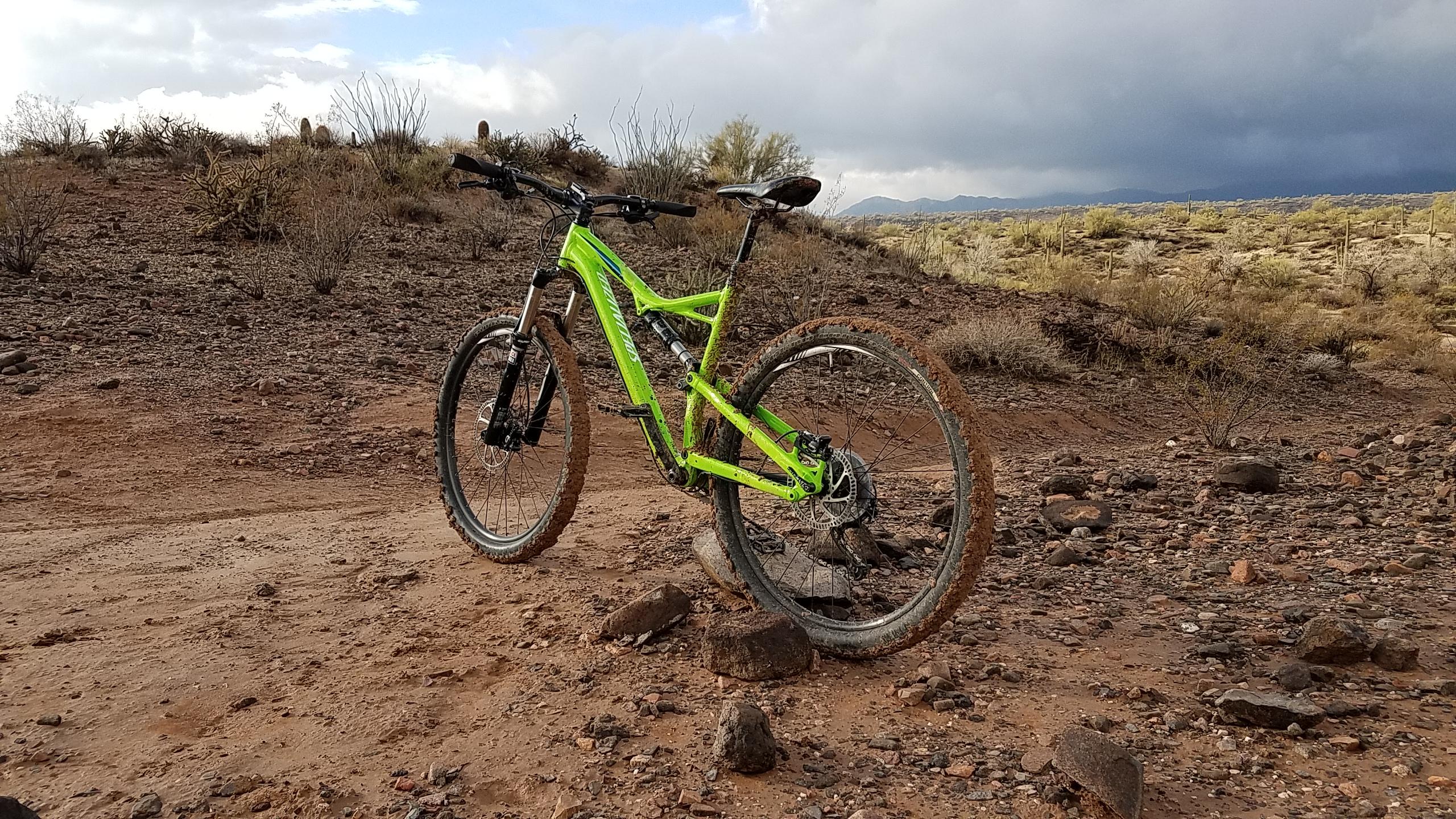 A bright green mountain bike rests on a rocky dirt trail with scattered vegetation in the background. The bike has mud on its tires, suggesting recent use on a rugged terrain. The sky is overcast, creating a dramatic atmosphere over the landscape. McDowell Mountain Park mountain bike trail.