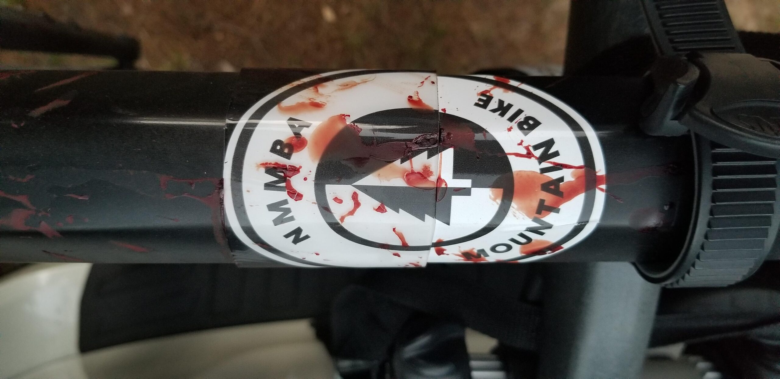 Niner Air 9 RDO: A close-up photo of a mountain bike frame showing a logo sticker, partially obscured by red splatters resembling blood. The background is blurred, featuring natural elements like leaves and dirt.