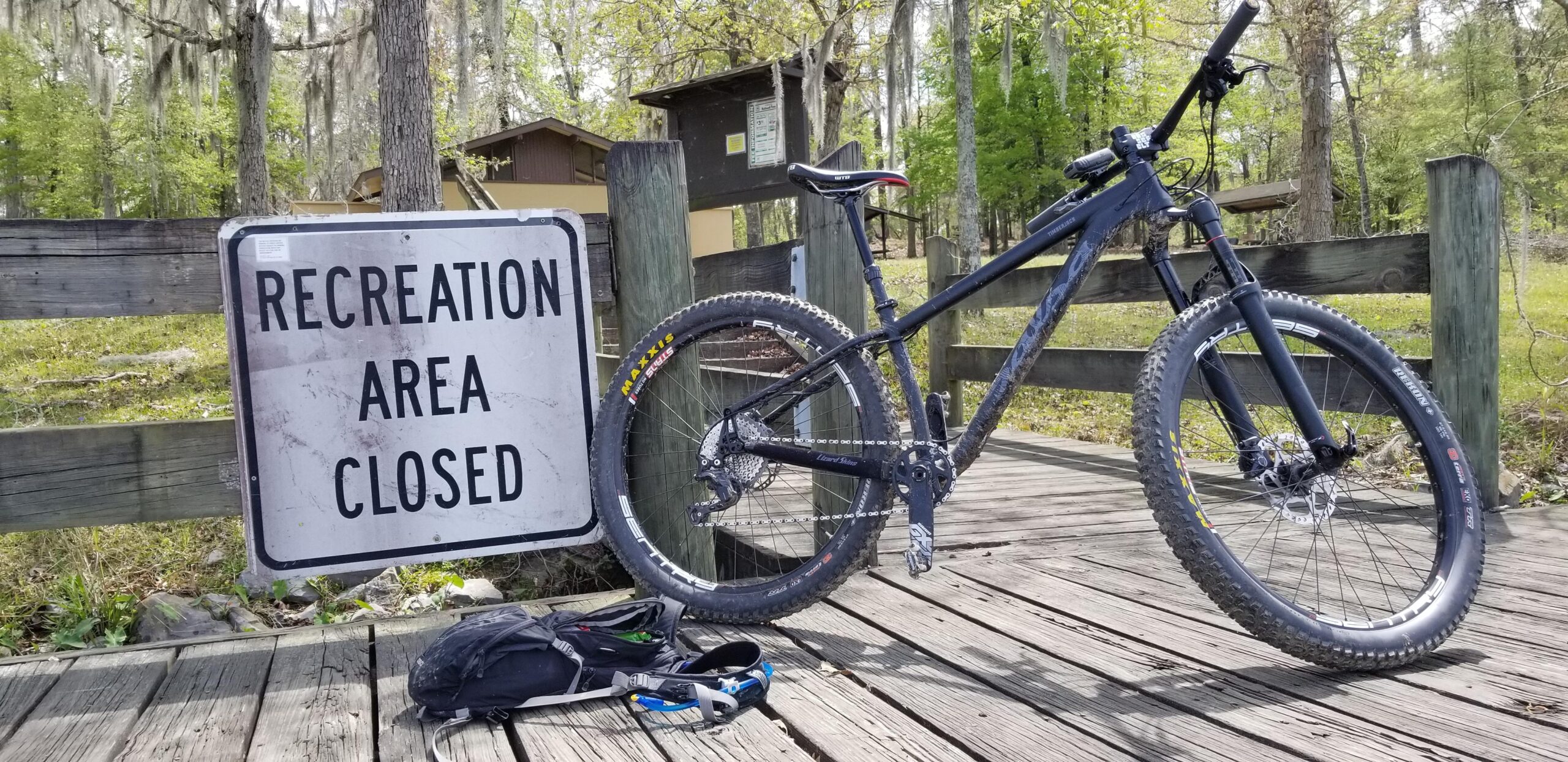 Salsa Timberjack: A black mountain bike leans against a sign that reads "RECREATION AREA CLOSED." The bike is positioned on a wooden bridge with a backpack lying on the ground nearby. In the background, trees with green foliage and a closed facility can be seen, indicating a natural park setting.