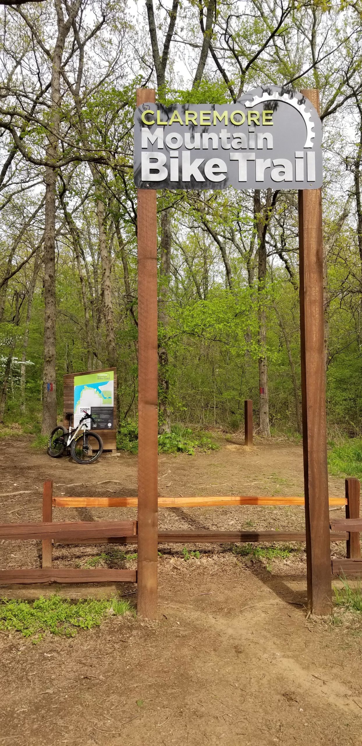 A sign marking the beginning of the Claremore Mountain Bike Trail, surrounded by trees and greenery. In the background, a map of the bike trail is displayed on a wooden structure, with a bicycle leaning against it. The area is natural and inviting, suitable for outdoor activities. Claremore mountain bike trail mountain bike trail.