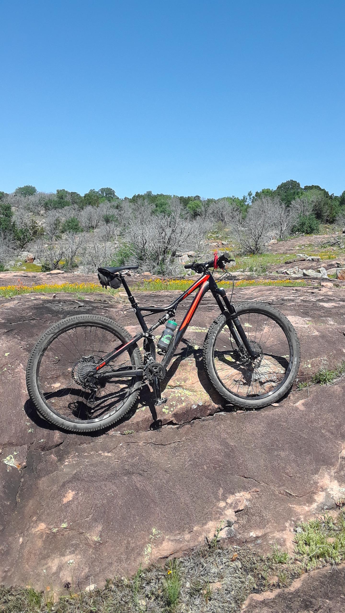 A mountain bike with black and orange accents is parked on a rocky surface, surrounded by sparse vegetation and patches of grass. The background features a clear blue sky and hills with a mix of green and dry trees. A water bottle is attached to the bike frame. Reveille Peak Ranch mountain bike trail.