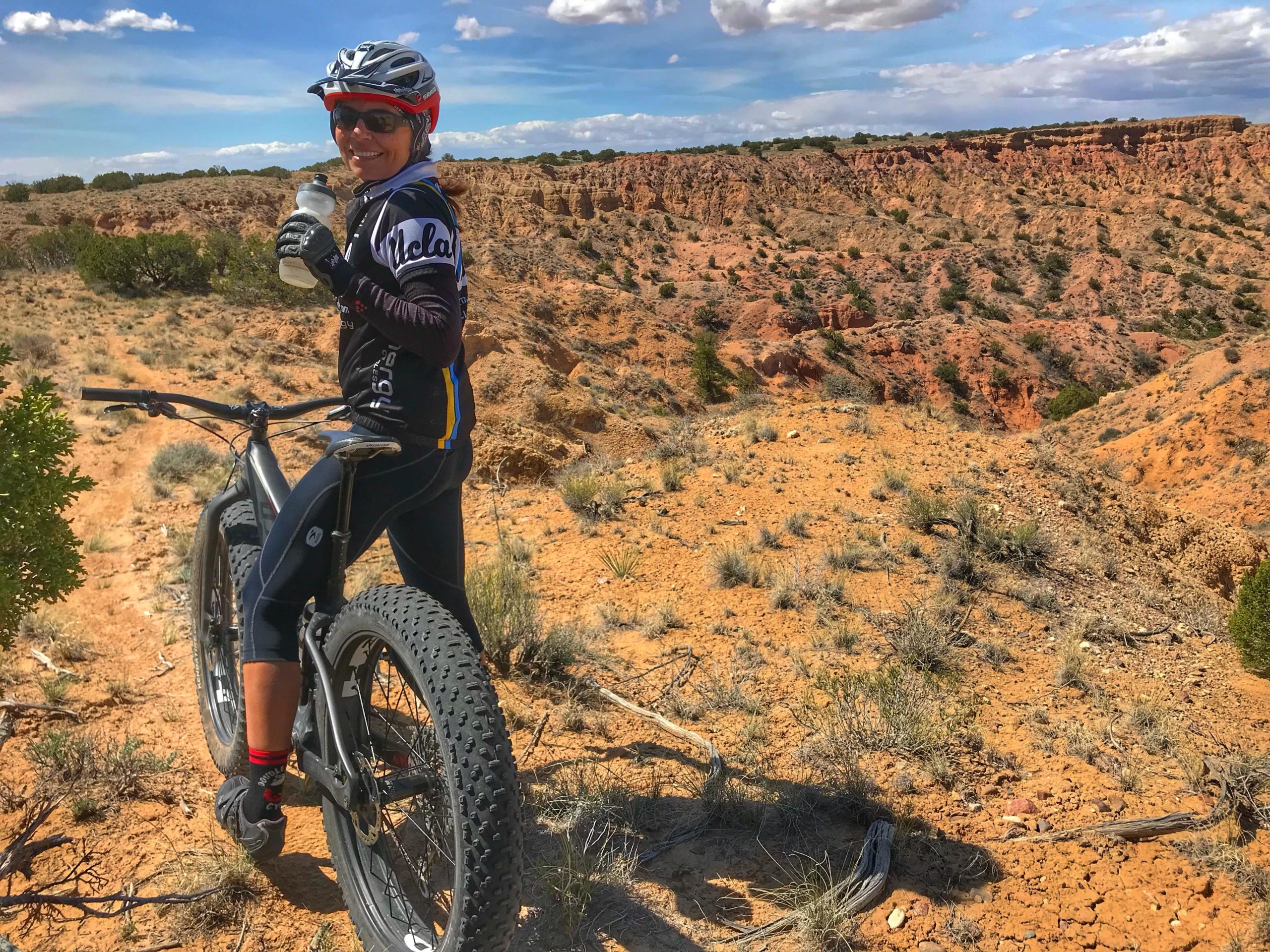 A mountain biker in a helmet and sunglasses smiles while holding a water bottle, positioned next to a fat bike on a sandy trail with rocky cliffs in the background. The scene is set against a bright blue sky with scattered clouds, showcasing a rugged and scenic landscape. Mariposa Fat Bike Trails mountain bike trail.