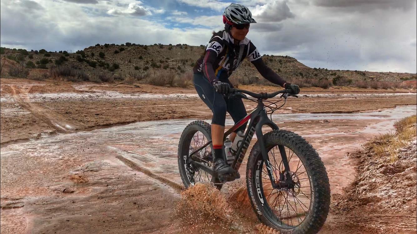 A person riding a fat bike through a muddy area, creating splashes, with a backdrop of hills and cloudy skies. The cyclist is wearing a helmet and cycling gear, showcasing an adventurous outdoor activity. Mariposa Fat Bike Trails mountain bike trail.
