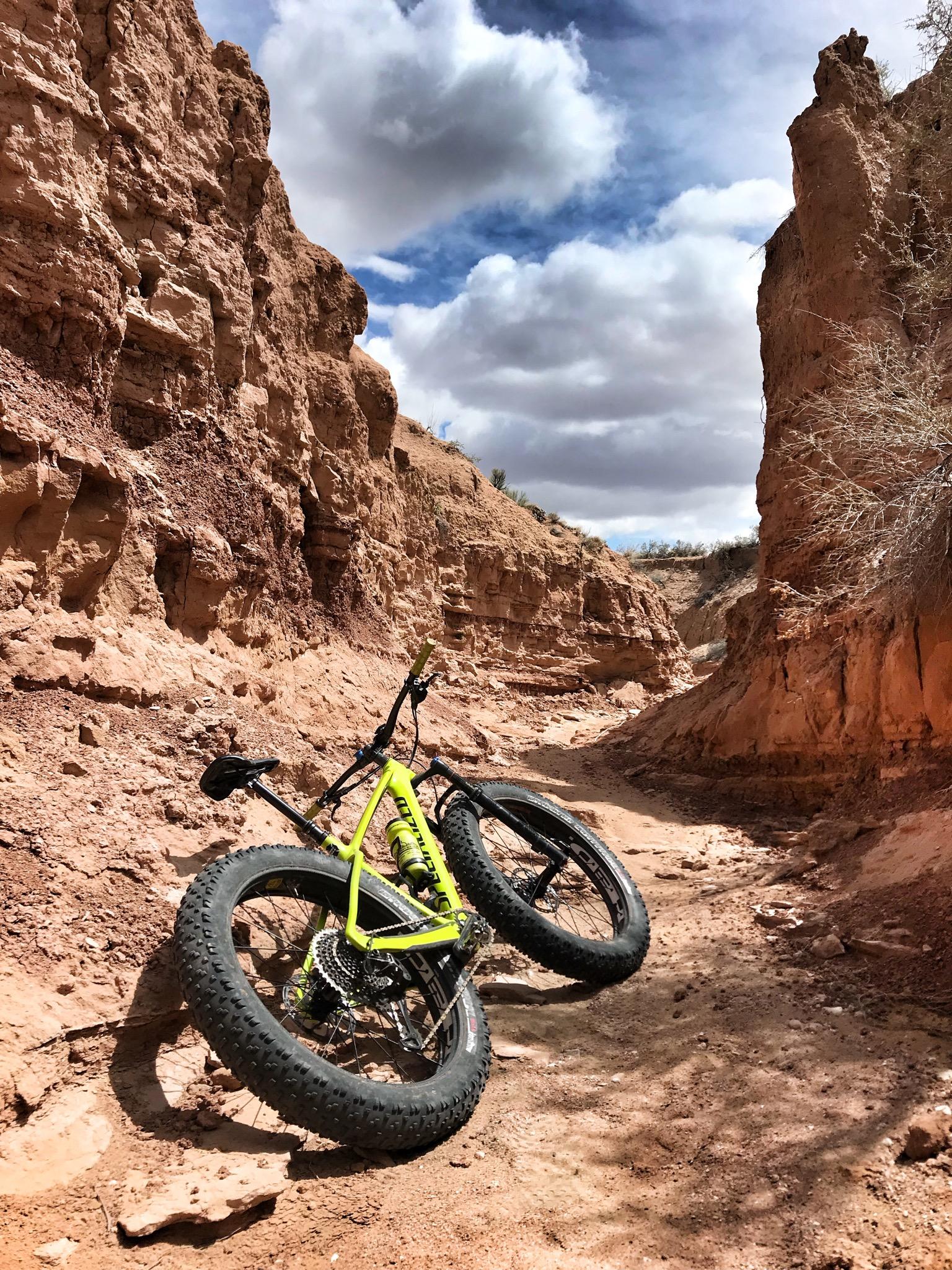 A bright yellow fat bike rests on a rugged dirt path surrounded by steep, reddish-brown canyon walls under a partly cloudy sky. The bike, equipped with wide tires suitable for off-road terrain, is positioned on its side, with the landscape showcasing natural erosion patterns and sparse vegetation. Mariposa Fat Bike Trails mountain bike trail.