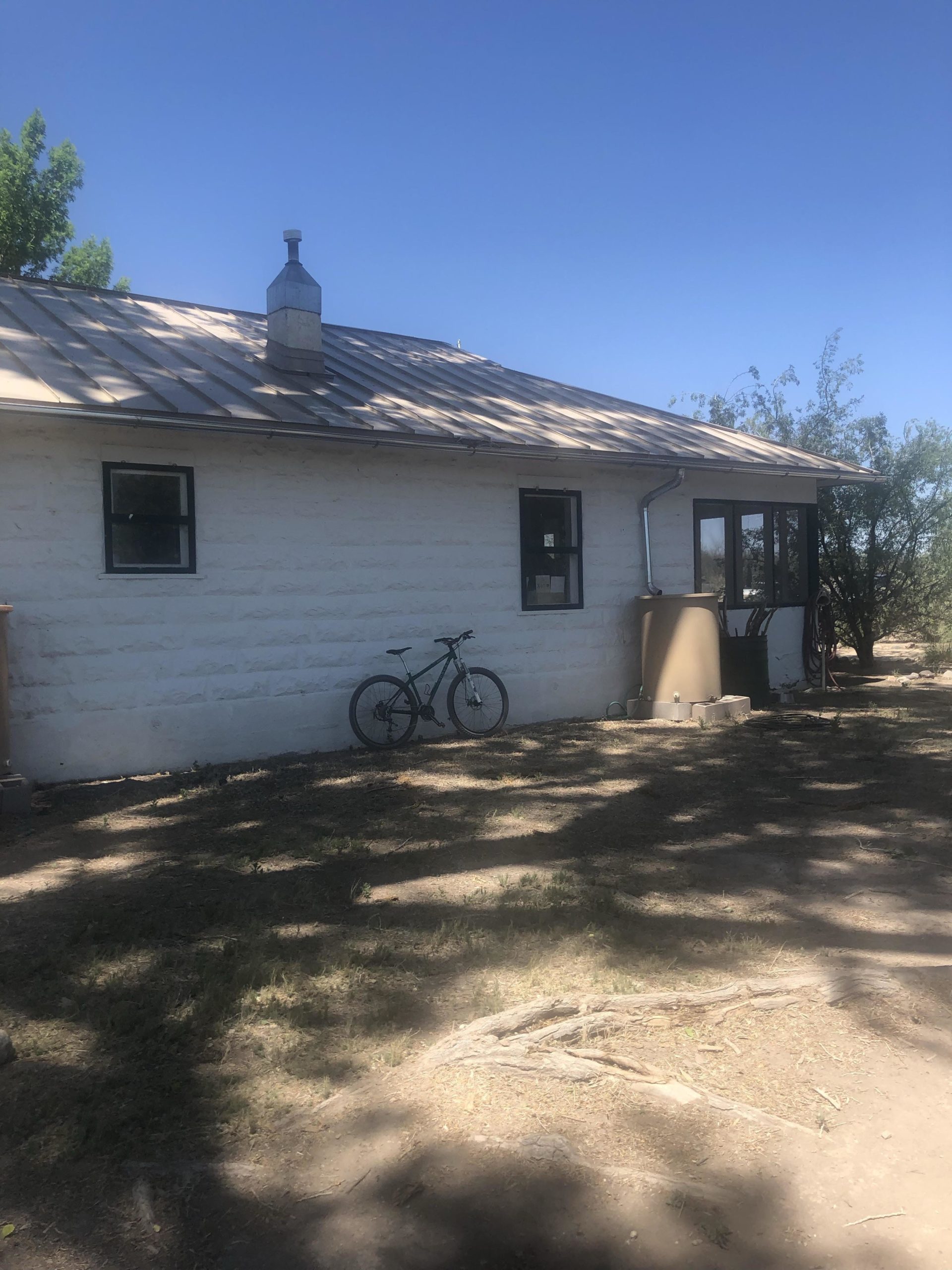A side view of a small, white bungalow with a metal roof, featuring a chimney. Leaning against the wall is a mountain bike, and the surrounding area is flat with sparse grass and some tree shade. The sky is clear and blue. Hereford To Waters Road Trailhead mountain bike trail.