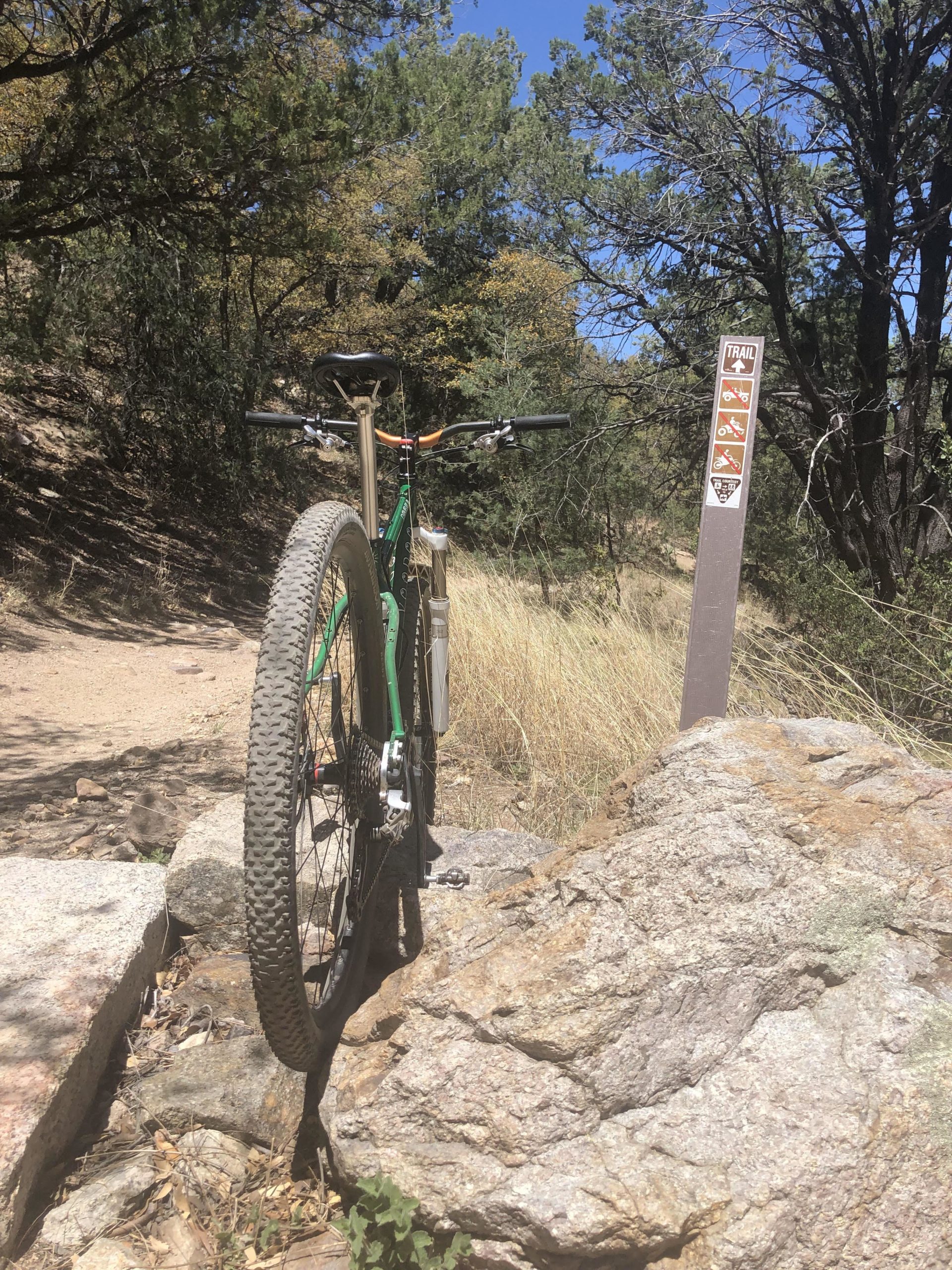 A mountain bike leaning against a rock on a dirt trail surrounded by trees and grass, with a trail sign featuring various symbols for trail use in the background. Brown Canyon mountain bike trail.