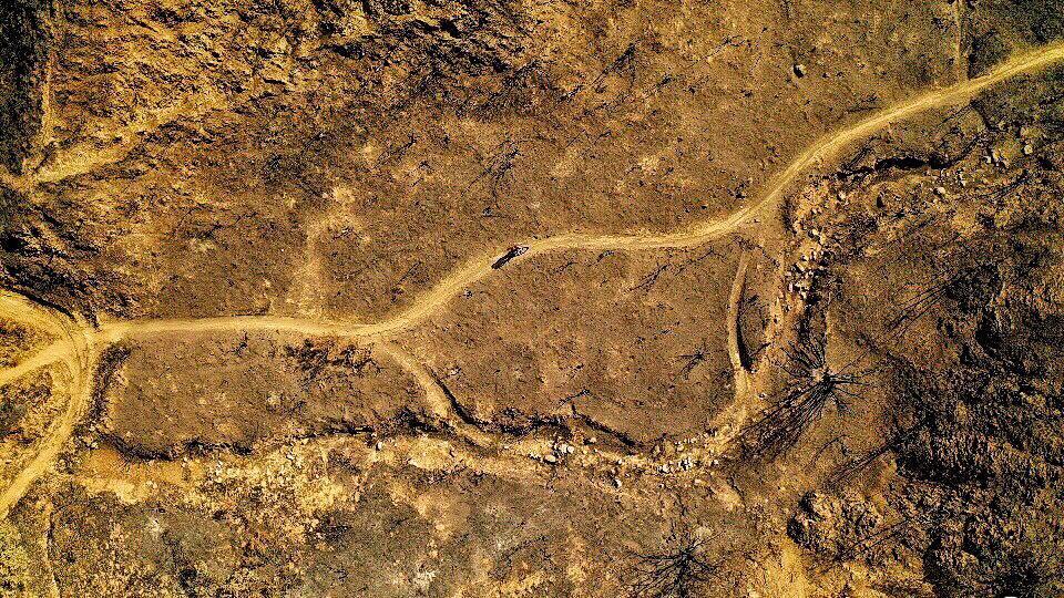 Giant Anthem SX 27.5: Aerial view of a barren landscape with a winding dirt path cutting through dry, sparse terrain. The ground appears cracked and brown, with patches of burnt vegetation and some tree stumps visible. The scene depicts the impact of drought or a wildfire.