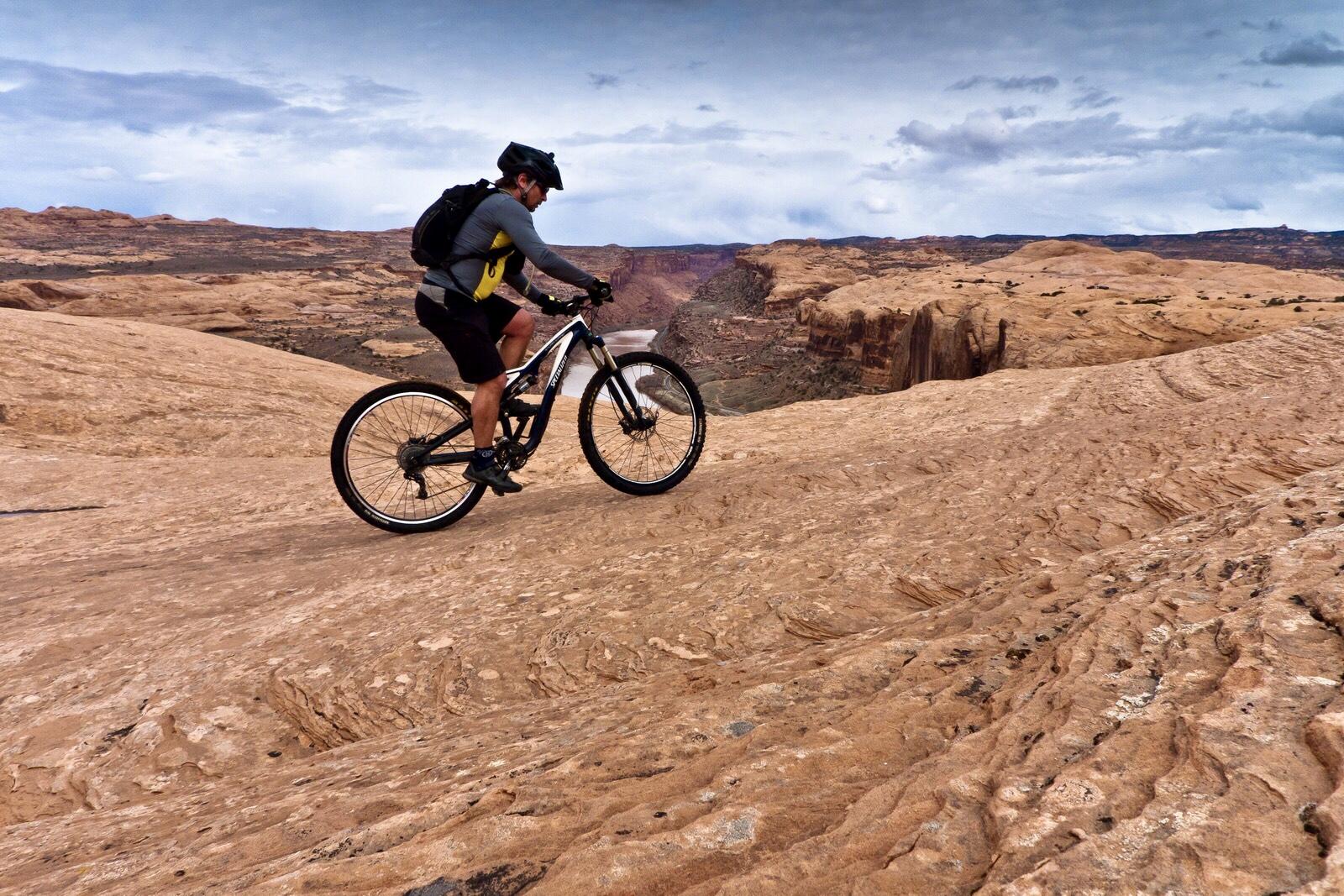 A mountain biker navigates a rocky terrain with dramatic cliffs and canyons in the background, under a cloudy sky. The cyclist wears a helmet and a backpack, showcasing a blend of adventure and nature. Slickrock mountain bike trail.