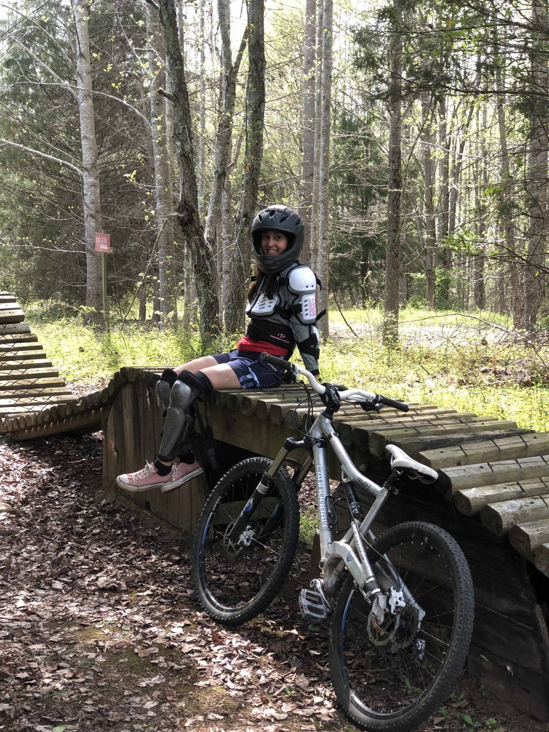 A mountain biker wearing protective gear, including a helmet and knee pads, sits on a wooden ramp in a forested area. A mountain bike rests beside her on the ramp, and the ground is covered with fallen leaves. Lush greenery and trees surround the scene, indicating a sunny day. Angler's Ridge mountain bike trail.