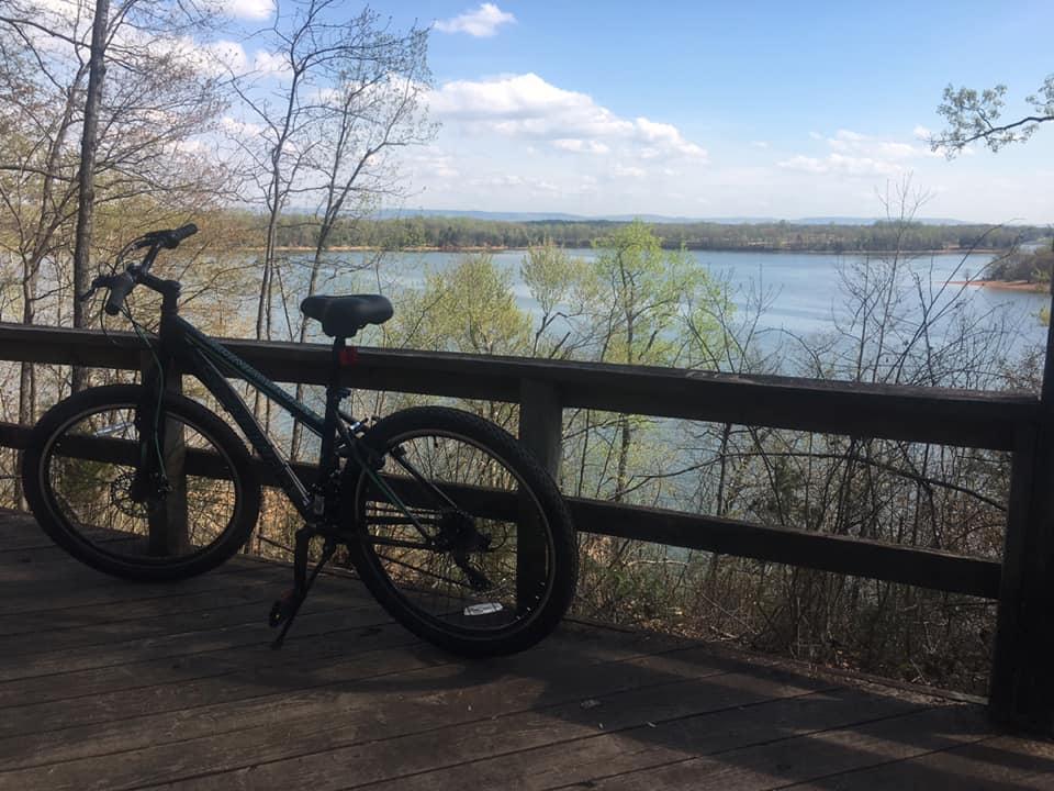 A bicycle is parked on a wooden deck overlooking a serene lake surrounded by trees and hills. The sky is partly cloudy, and the scene conveys a peaceful outdoor atmosphere. Tims Ford State Park mountain bike trail.