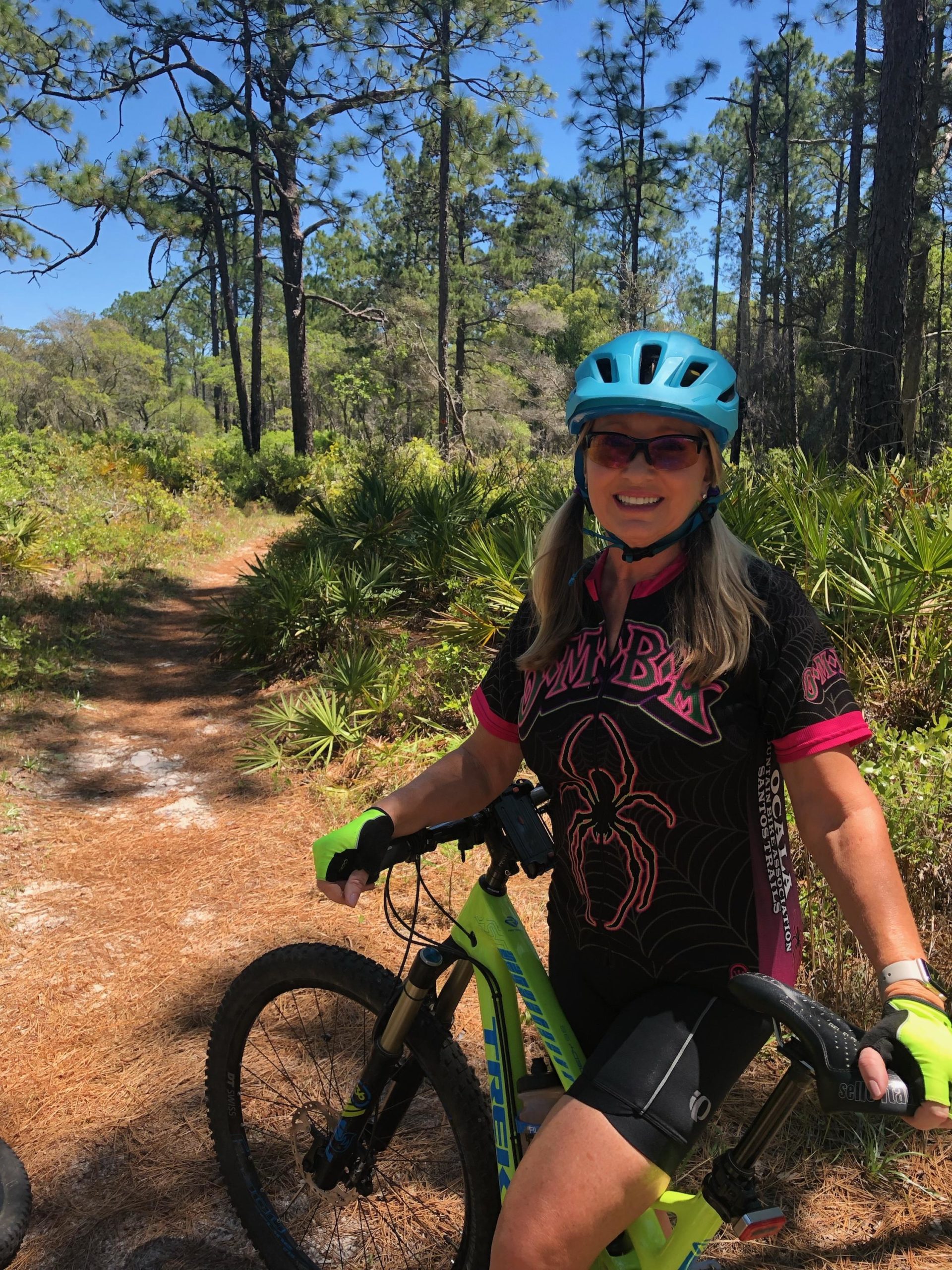 A woman wearing a turquoise helmet and sunglasses stands beside a green mountain bike on a dirt trail surrounded by tall trees and lush greenery. She is smiling and dressed in a black and pink cycling jersey, with gloves and shorts, ready for a ride in a sunny outdoor setting. Longleaf Pine Greenway Trail mountain bike trail.