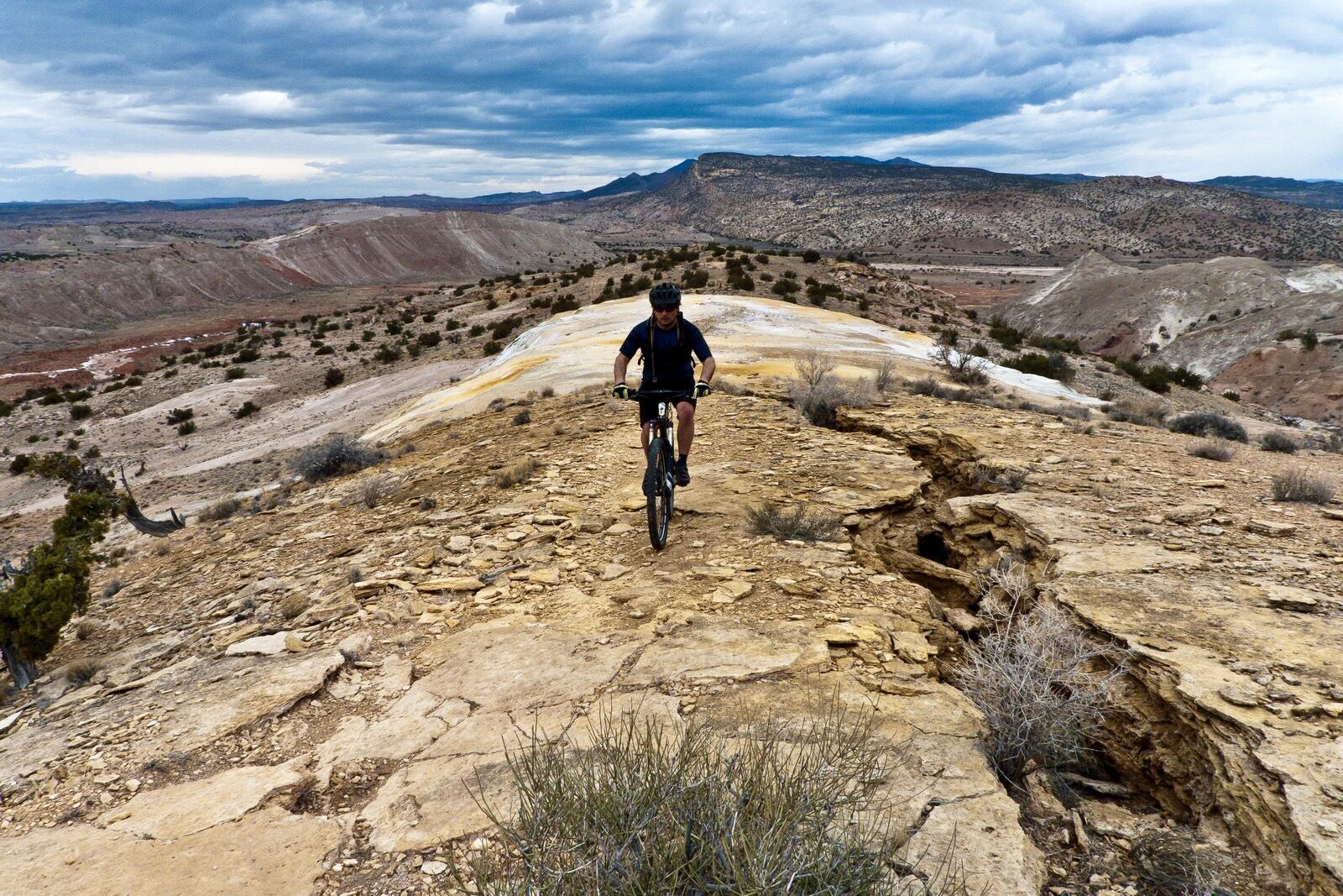 A mountain biker navigates a rocky trail with a scenic desert landscape in the background, featuring rugged terrain and cloudy skies. White Ridge Bike Trails mountain bike trail.