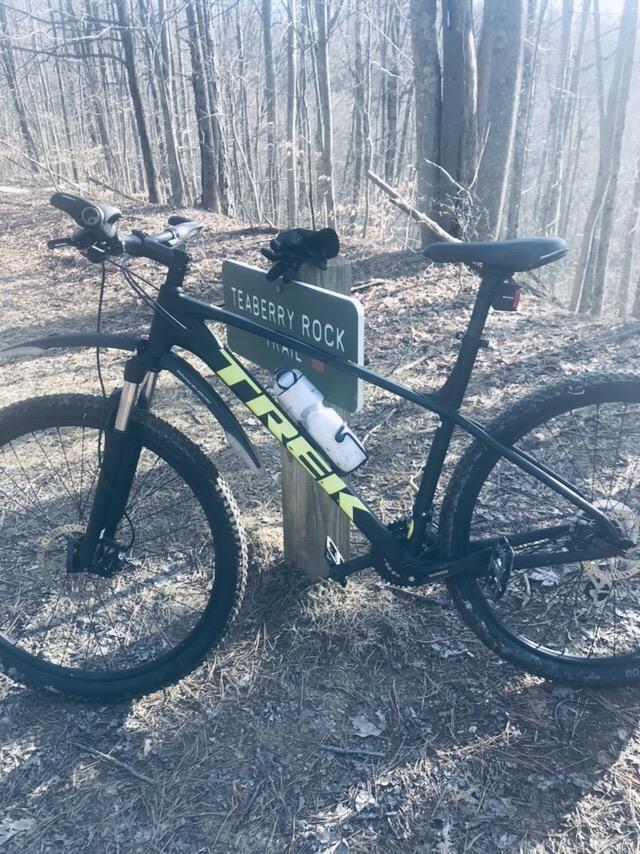 Trek Marlin 6: A black and green mountain bike is leaning against a trail sign that reads "Teaberry Rock Trail." The bike features a water bottle holder along the frame and is surrounded by a wooded area with leafless trees, suggesting an outdoor adventure setting.