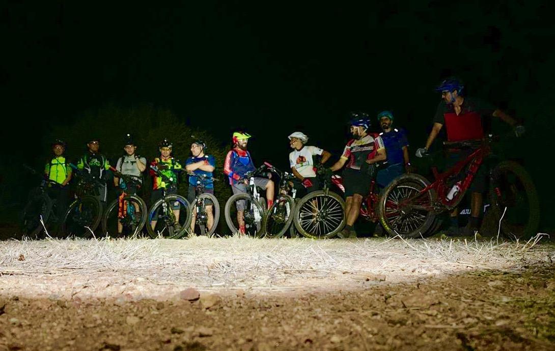 Group of eleven mountain bikers gathered at night, illuminated by a light source, with various colorful cycling gear and helmets. They are positioned with their bikes on a dirt path surrounded by dark vegetation. Jubileo mountain bike trail.