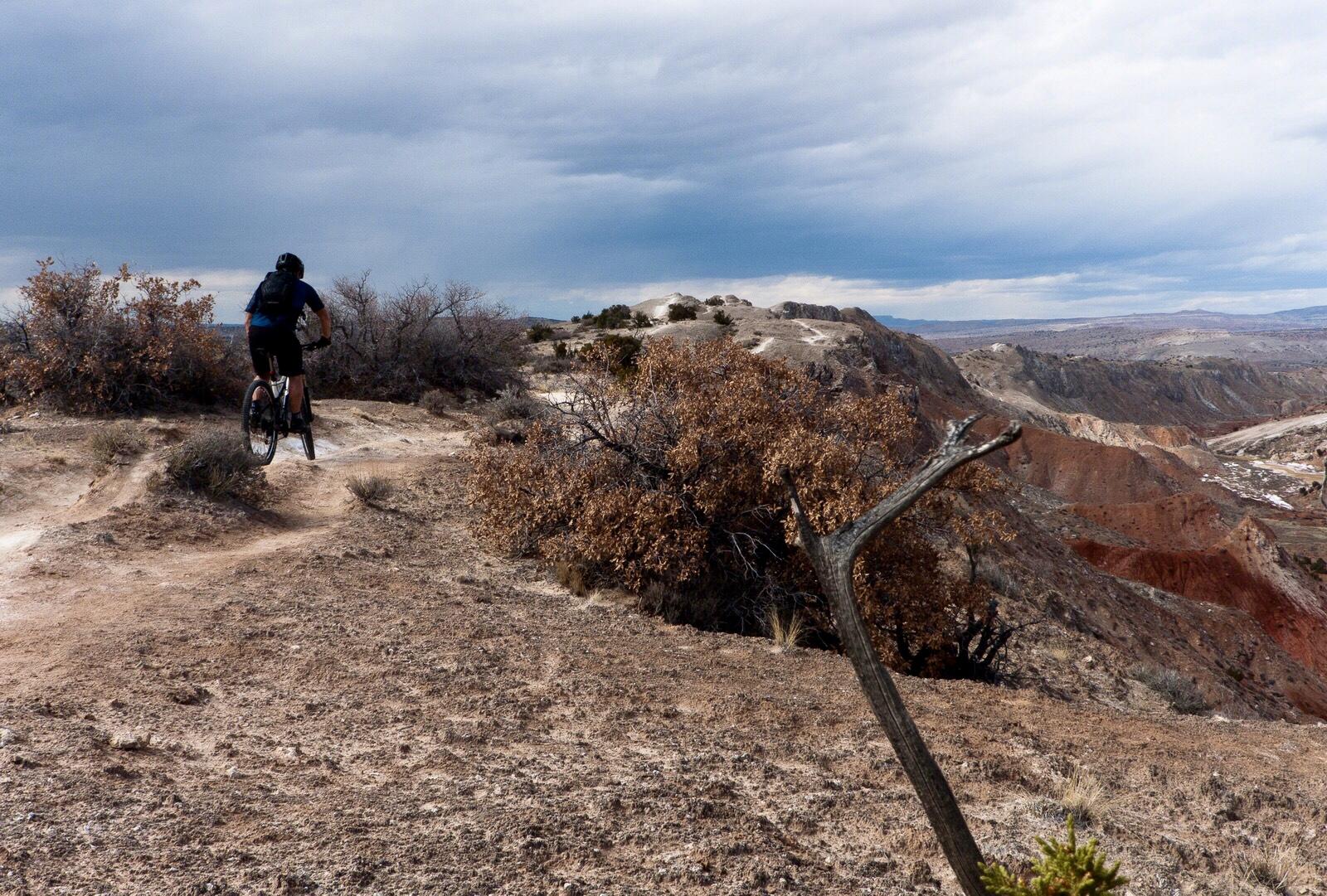 A mountain biker riding along a dirt trail surrounded by sparse vegetation, with a rocky landscape and cloudy skies in the background. The scene captures the rugged beauty of the terrain. White Ridge Bike Trails mountain bike trail.