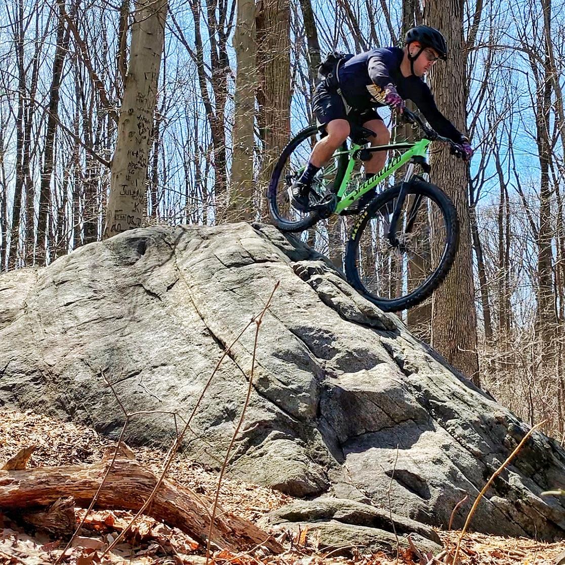 A cyclist mid-jump off a large rock formation in a forested area, surrounded by bare trees and scattered leaves. The cyclist is wearing a helmet, dark clothing, and riding a green mountain bike. Brandywine State Park mountain bike trail.