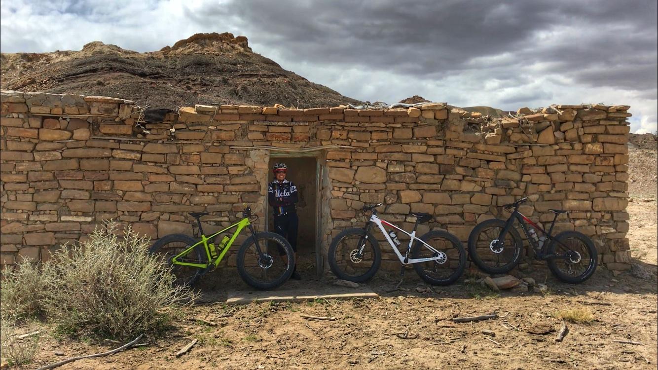 A person stands in the doorway of a rustic stone building, smiling. Three mountain bikes are propped up beside the building: a green bike on the left, a white bike in the center, and a black bike on the right. The landscape is arid, with sparse vegetation and a cloudy sky overhead. Mariposa Fat Bike Trails mountain bike trail.