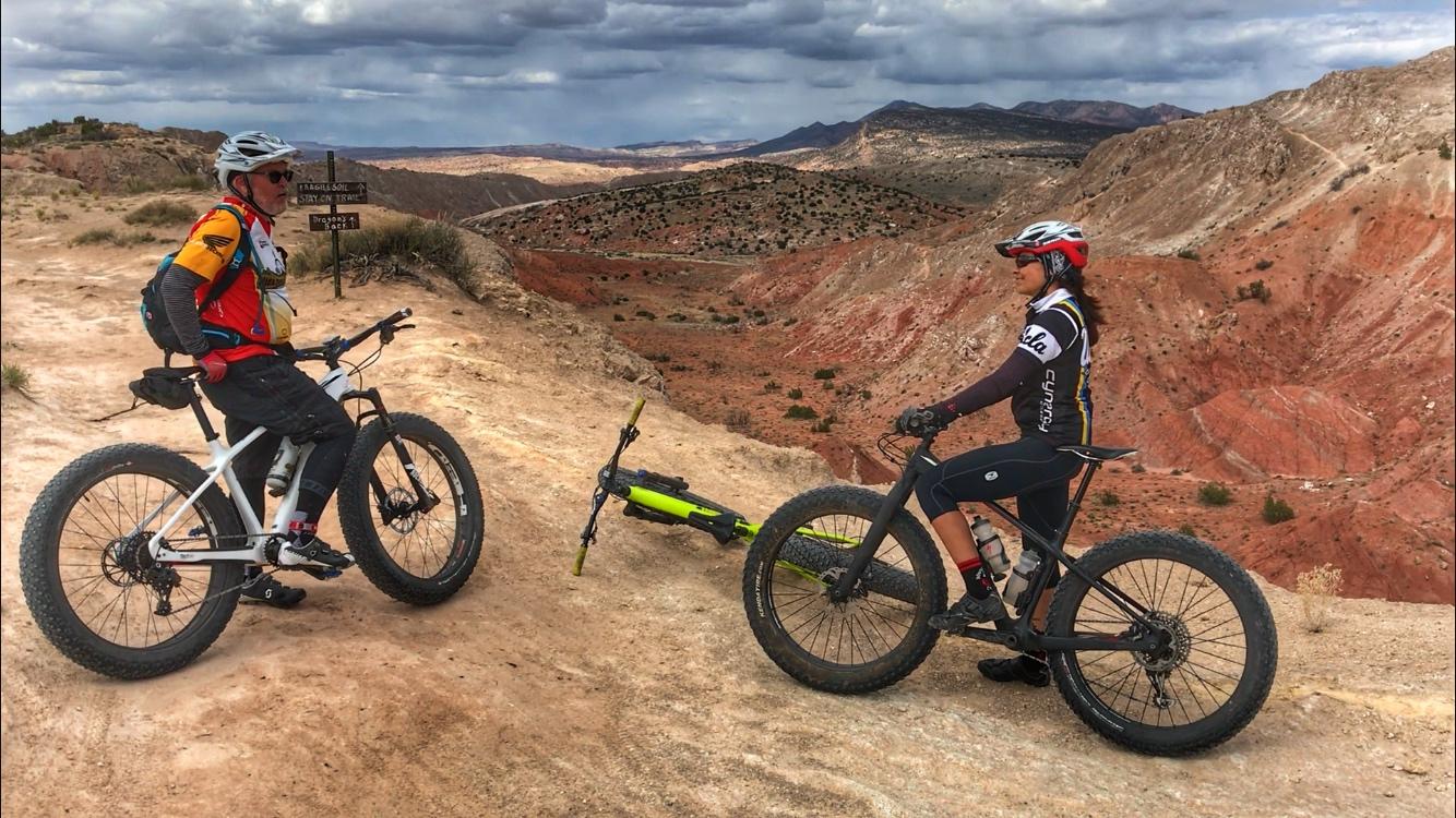 Two mountain bikers pause on a rocky trail overlooking a vast, colorful landscape. One cyclist, dressed in a bright red and blue jersey, stands with a white bike, while the other, wearing a black and white jersey, is seated on a black bike. The terrain features red and beige hues, with distant hills under a cloudy sky. A signpost in the background alerts hikers to stay on the trail. Mariposa Fat Bike Trails mountain bike trail.