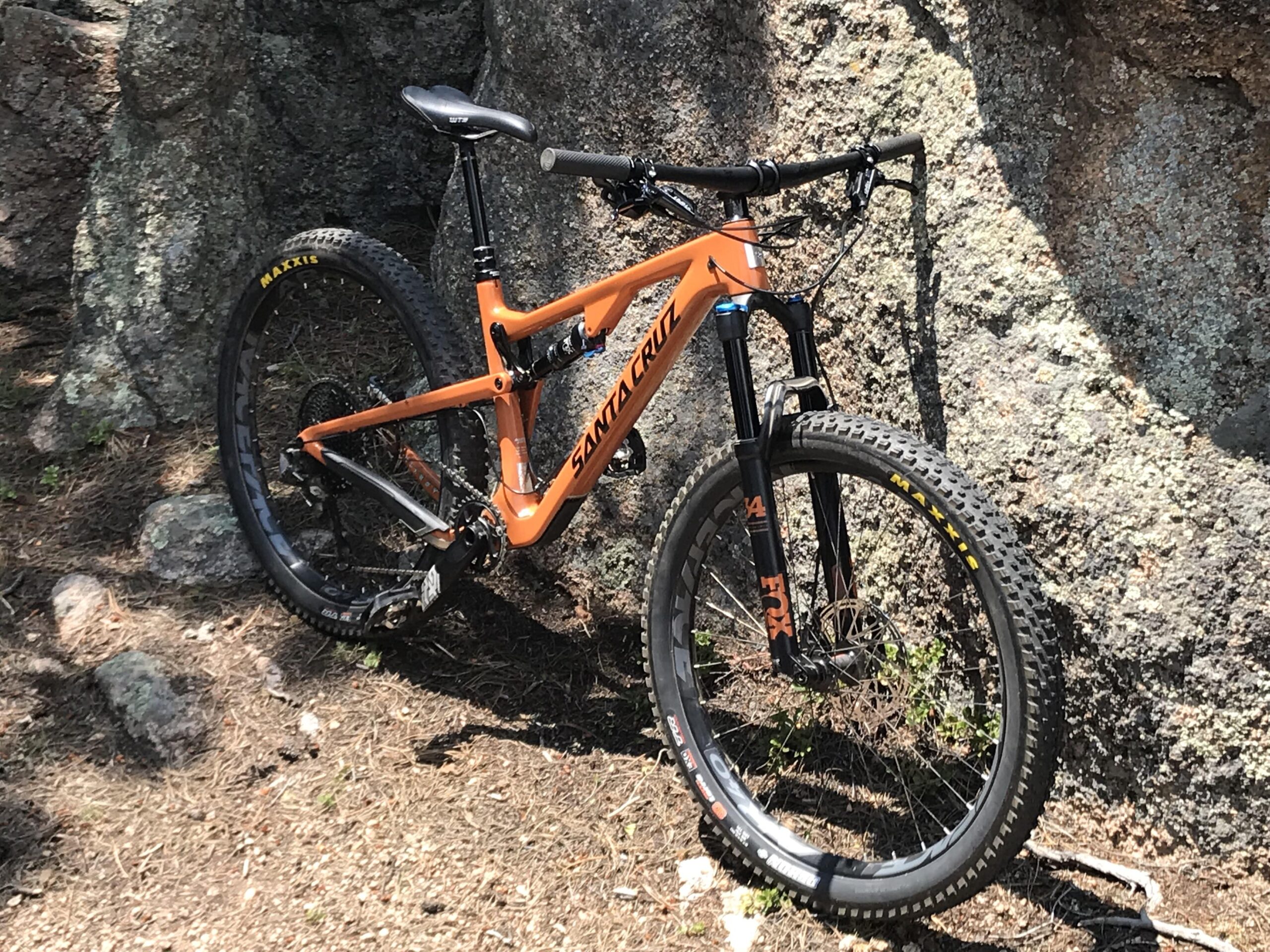 Santa Cruz Tallboy Carbon: A bright orange mountain bike leaning against a rocky surface, surrounded by dirt and pine needles. The bike features wide tires and a robust frame, designed for off-road riding.