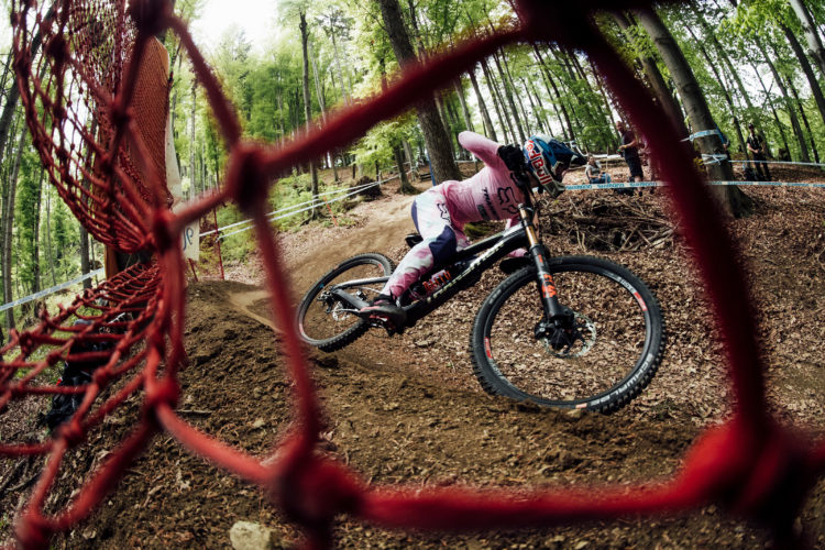 A mountain biker in a pink and white jersey leans into a turn on a dirt track, framed by a protective red netting. Surrounding trees create a natural backdrop, while a few spectators watch in the distance. The scene captures the excitement and intensity of downhill racing.