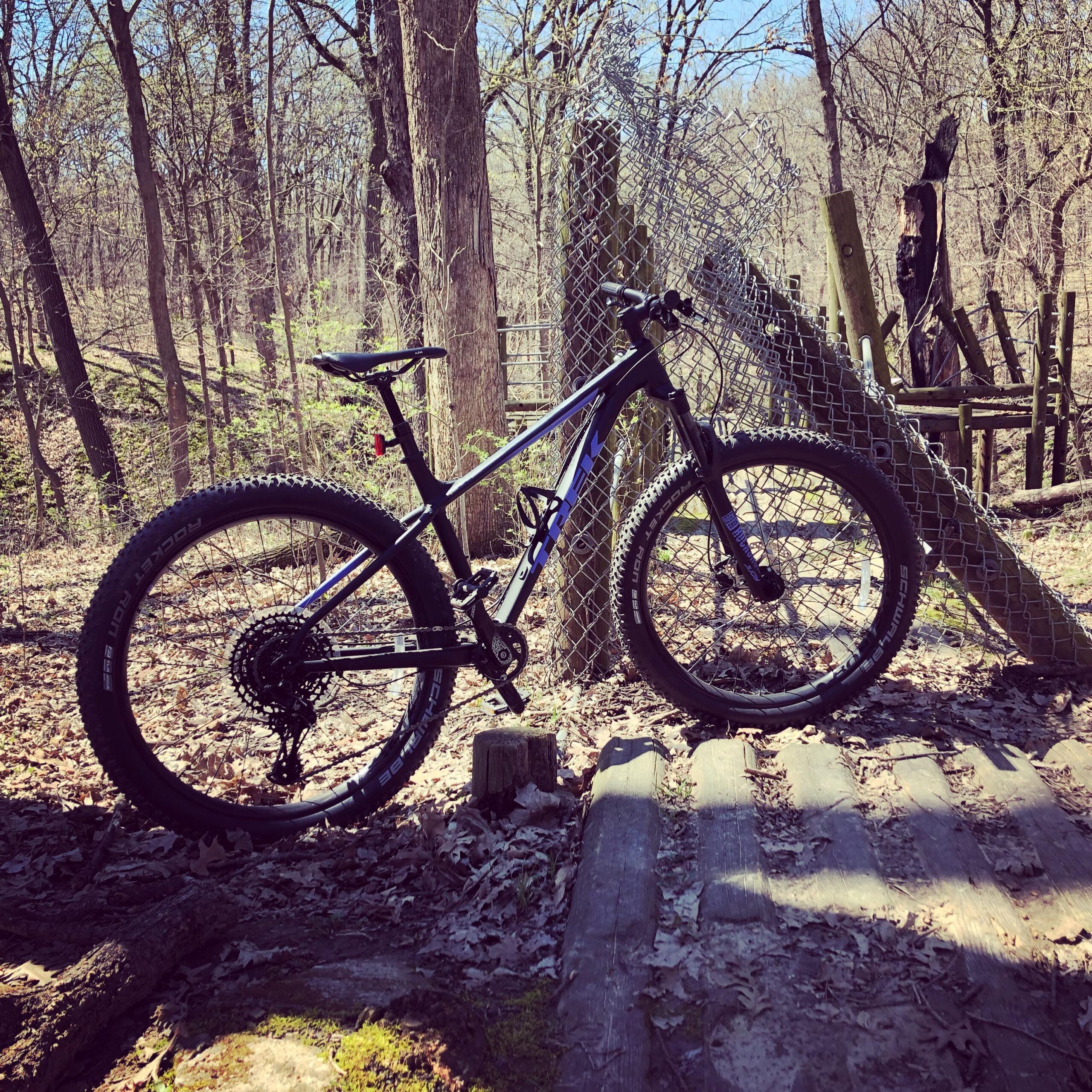 Trek Roscoe 8: A mountain bike parked on a wooden path in a forested area, surrounded by trees and fallen leaves, with a chain-link fence and wooden structures in the background. The scene is bright and inviting, indicating a sunny day.