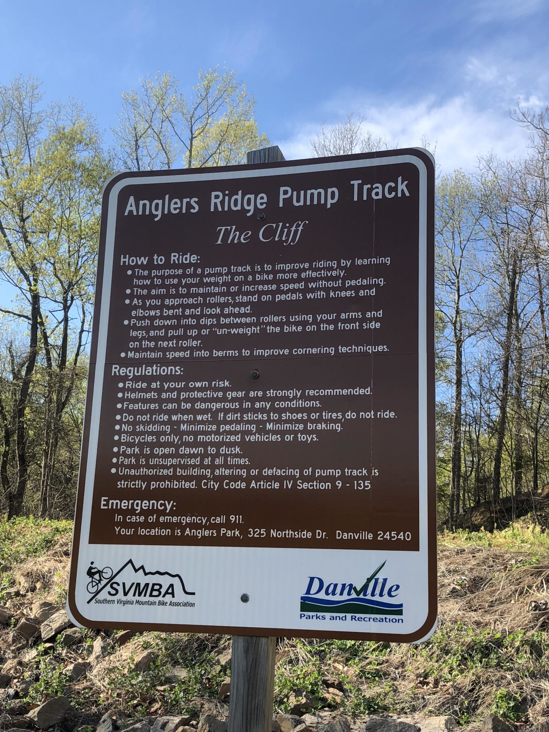 A sign at Anglers Ridge Pump Track titled "The Cliff," providing instructions and safety guidelines for riding. The sign outlines how to effectively use the pump track, emphasizing techniques for maintaining speed and improving biking skills. It also includes regulations such as riding at one's own risk, the necessity of wearing helmets, and reminders about park usage. The background features trees in early spring. Angler's Ridge mountain bike trail.